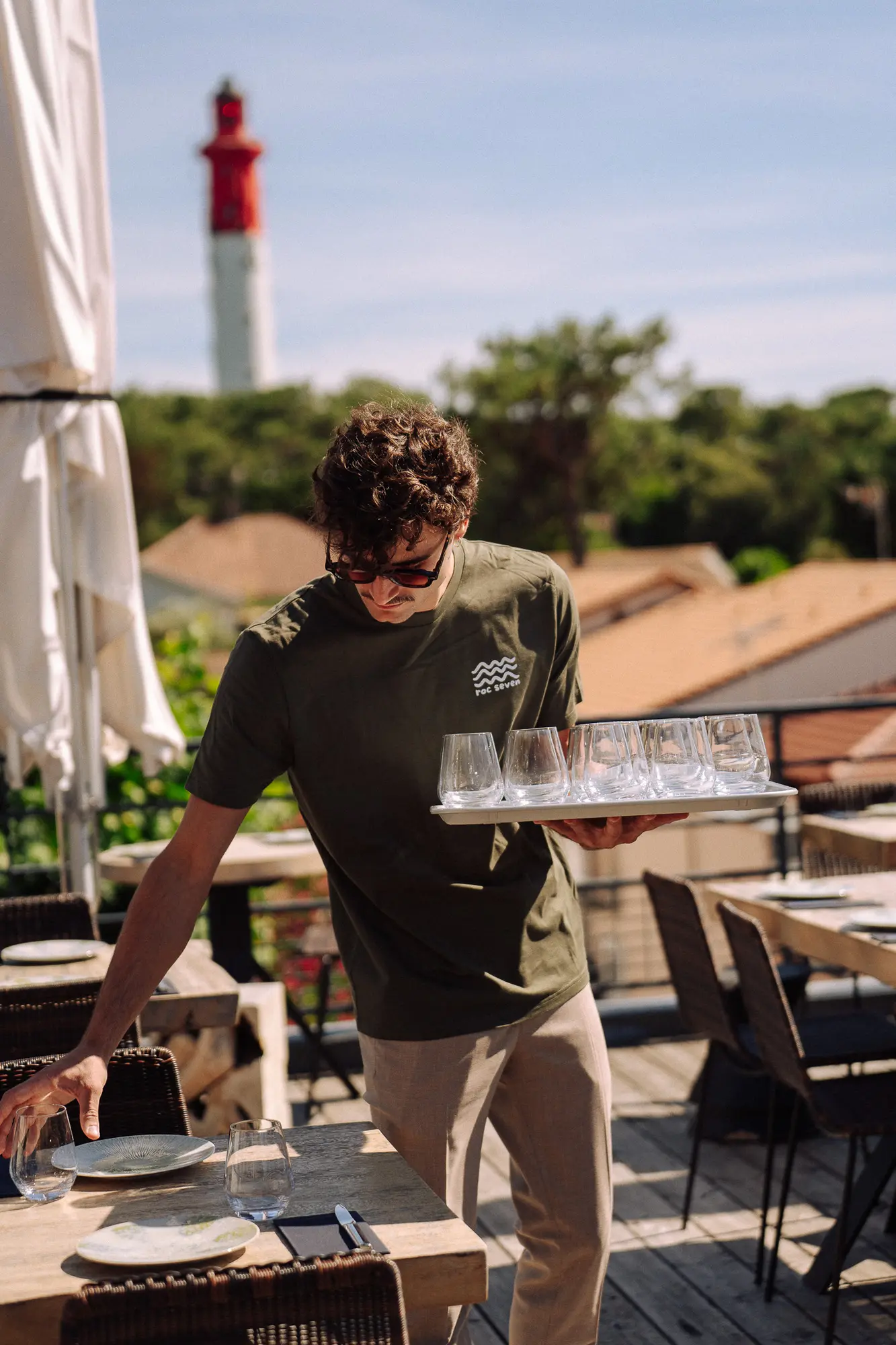 Un serveur portant un t-shirt vert avec le logo "roc seven" sert des verres sur une terrasse ensoleillée d'un restaurant avec une vue sur un phare rouge et blanc en arrière-plan.