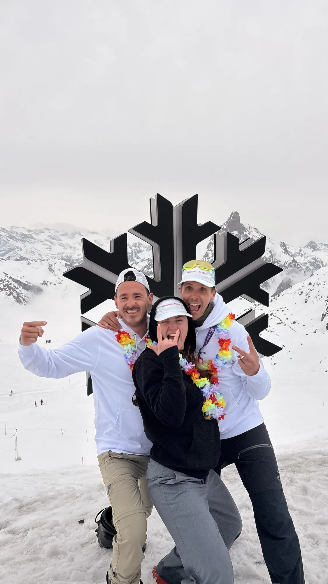 Trois amis posant joyeusement devant une grande sculpture en forme de flocon de neige noir sur un fond de montagnes enneigées, portant des vêtements d'hiver décontractés et des colliers de fleurs multicolores.