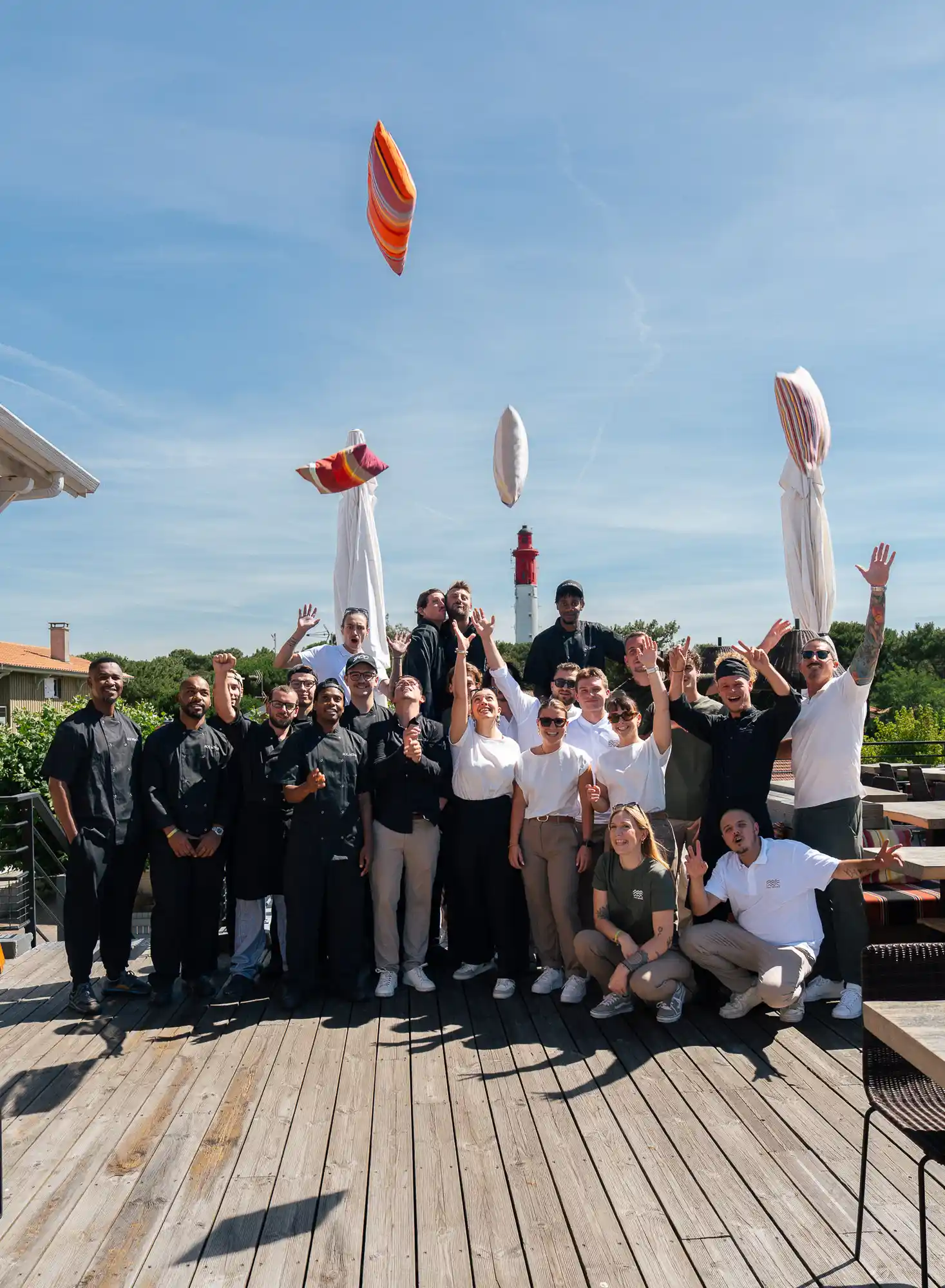 Groupe de jeunes adultes posant sur une terrasse en bois, certains lançant des coussins colorés en l'air, avec un phare en arrière-plan sous un ciel bleu clair.