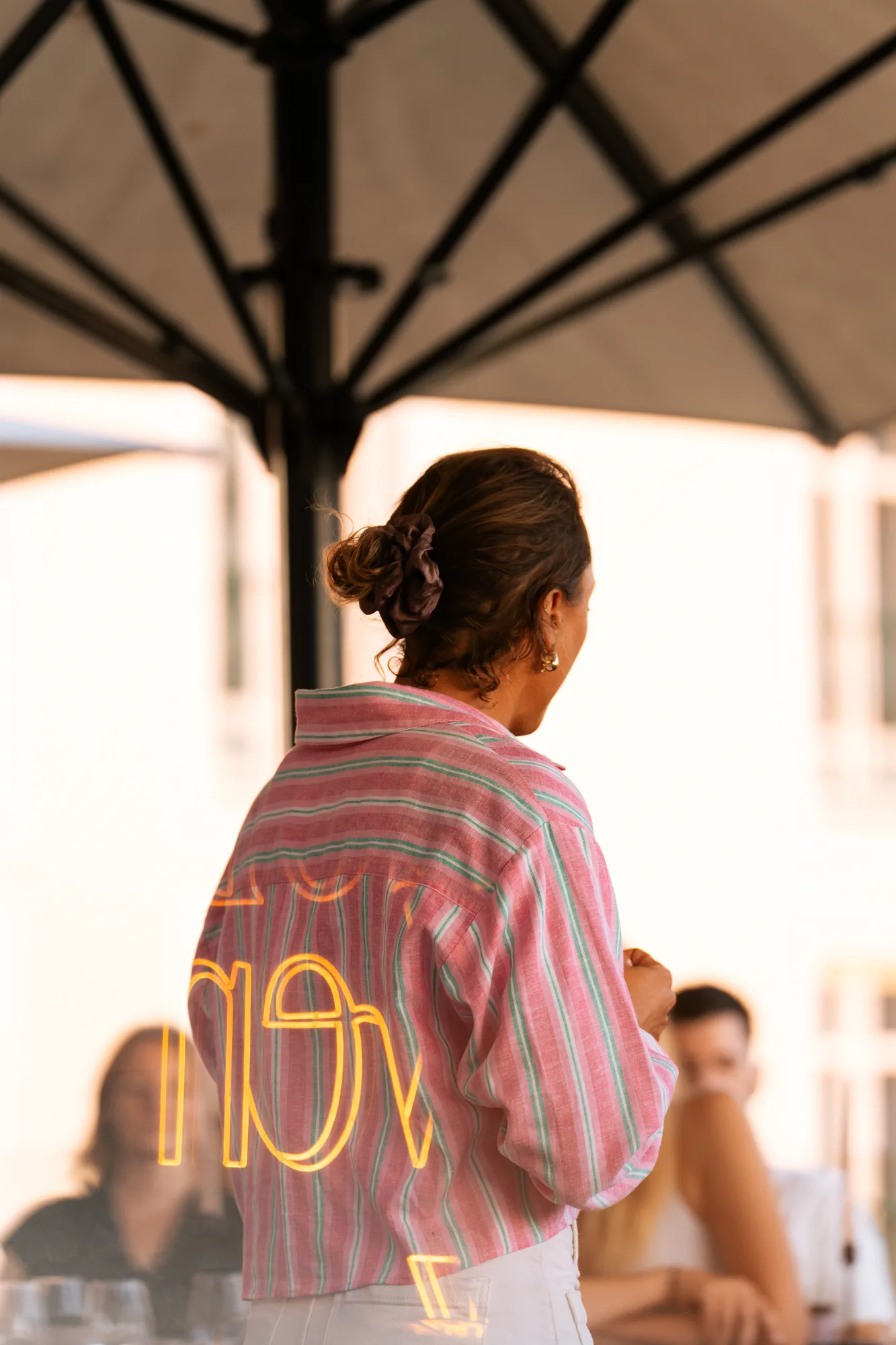 Femme de dos portant une chemise rayée rose et verte, les cheveux attachés avec un scrunchie, sous un parasol noir, avec des personnes floues en arrière-plan.