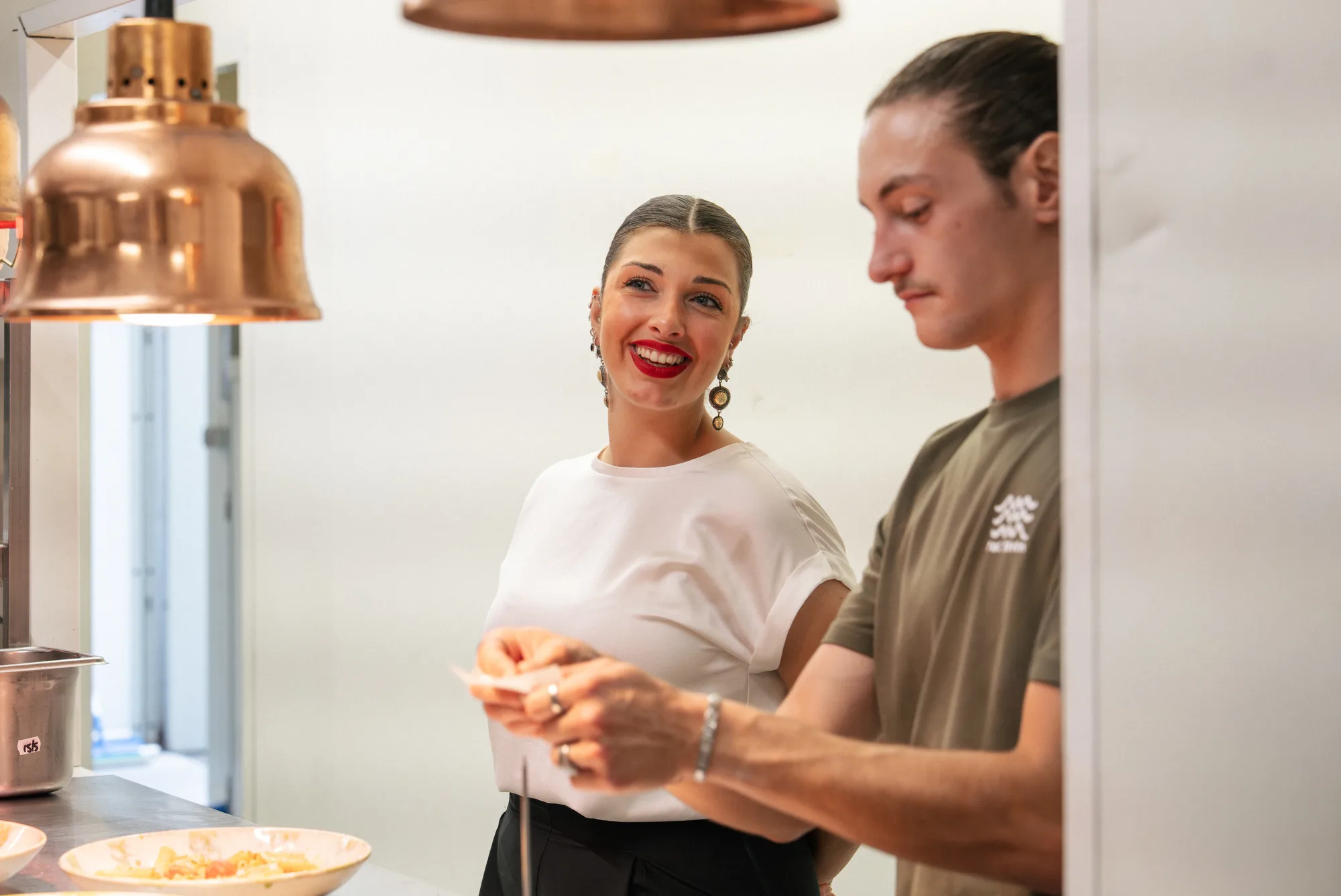 Une femme souriante avec du rouge à lèvres rouge et des boucles d'oreilles pendantes regarde un homme concentré tenant une note dans une cuisine professionnelle.
