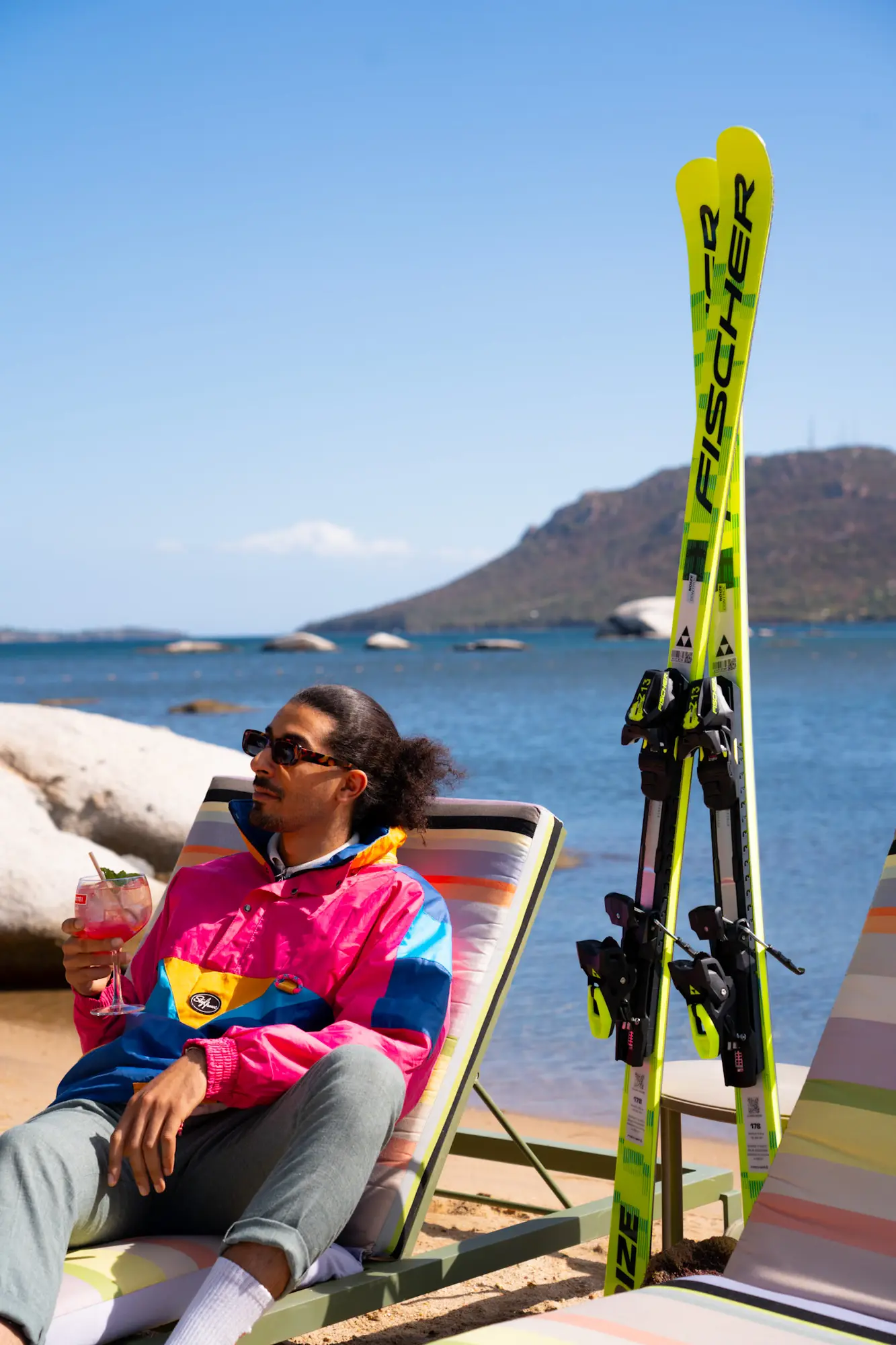Un homme avec une veste colorée assis sur une chaise longue à rayures sur une plage, tenant un verre de cocktail, avec des skis Fischer jaunes appuyés à côté, et un lac avec des rochers et une montagne en arrière-plan sous un ciel bleu clair.
