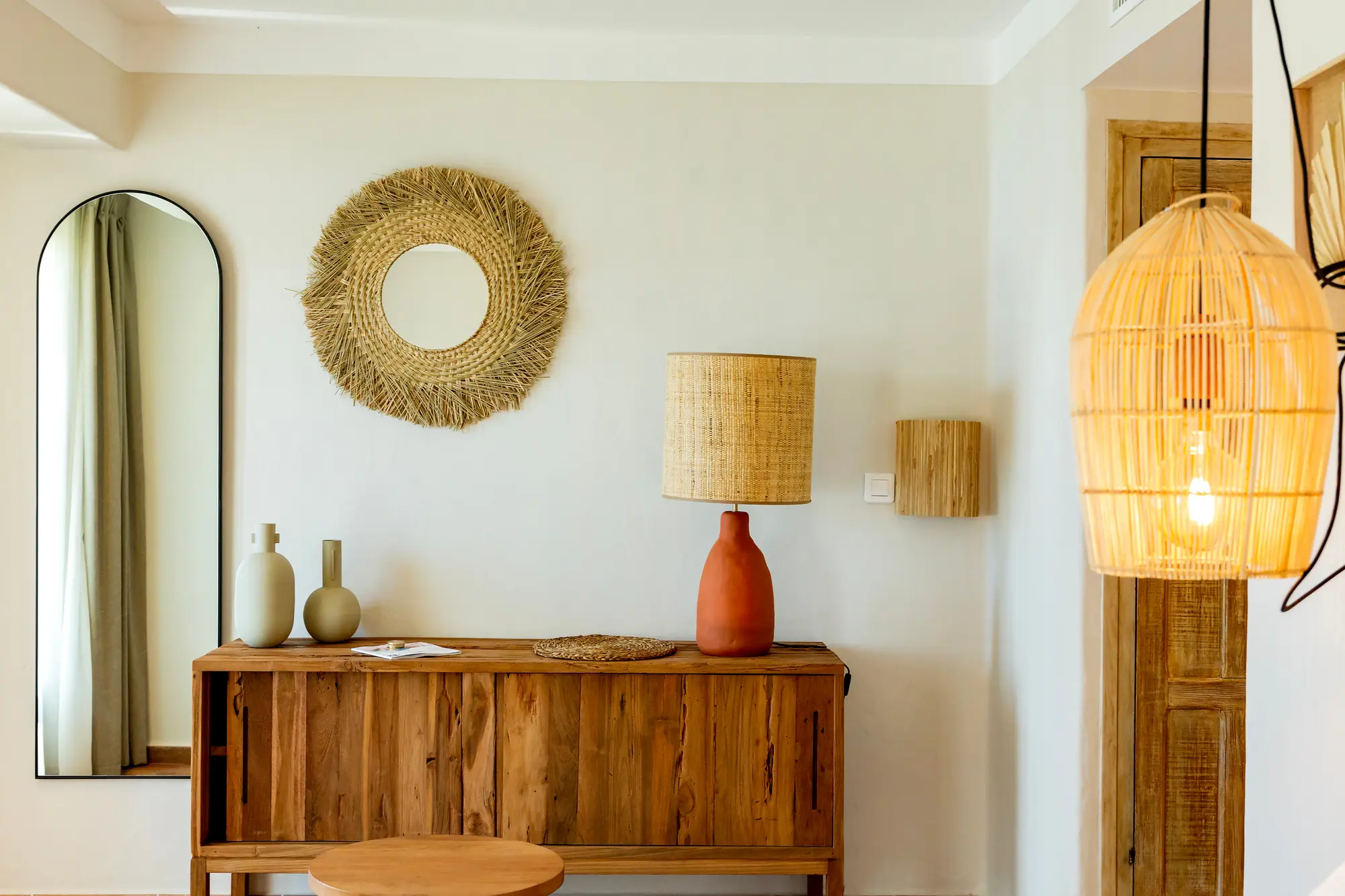 Intérieur avec meuble en bois naturel surmonté d'une lampe avec abat-jour en fibre tressée, deux vases de céramique et un miroir mural rond encadré de fibres naturelles, près d'un miroir mural vertical à cadre noir et d'une suspension lumineuse en rotin.