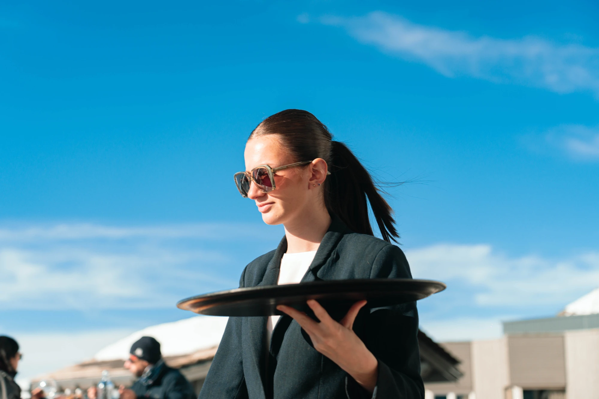 Serveuse portant un plateau vide, portant des lunettes de soleil et un blazer noir, avec un ciel bleu en arrière-plan.