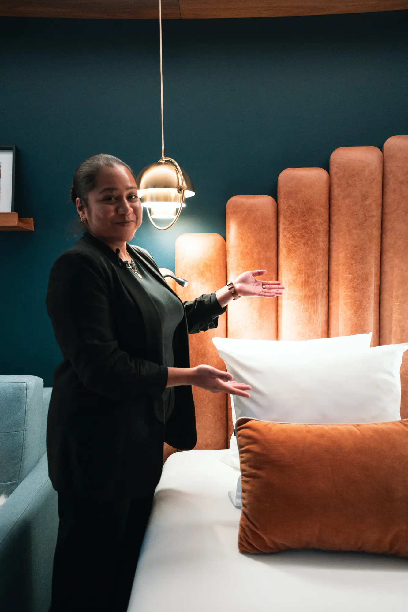 Femme en costume noir présentant un lit avec une tête de lit en cuir marron clair, des oreillers blancs et un coussin orange, dans une chambre avec un mur bleu foncé et une lampe suspendue dorée.