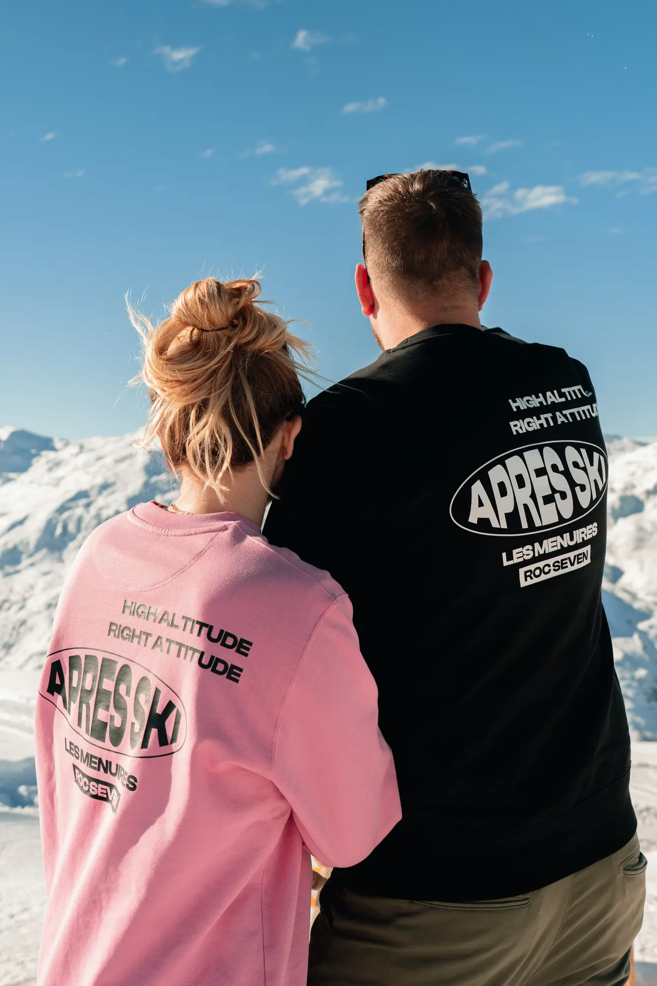 Un couple vu de dos porte des sweat-shirts avec les inscriptions "HIGH ALTITUDE RIGHT ATTITUDE APRES SKI LES MENUIRES ROC SEVEN" sur un fond de paysage enneigé et ciel bleu.