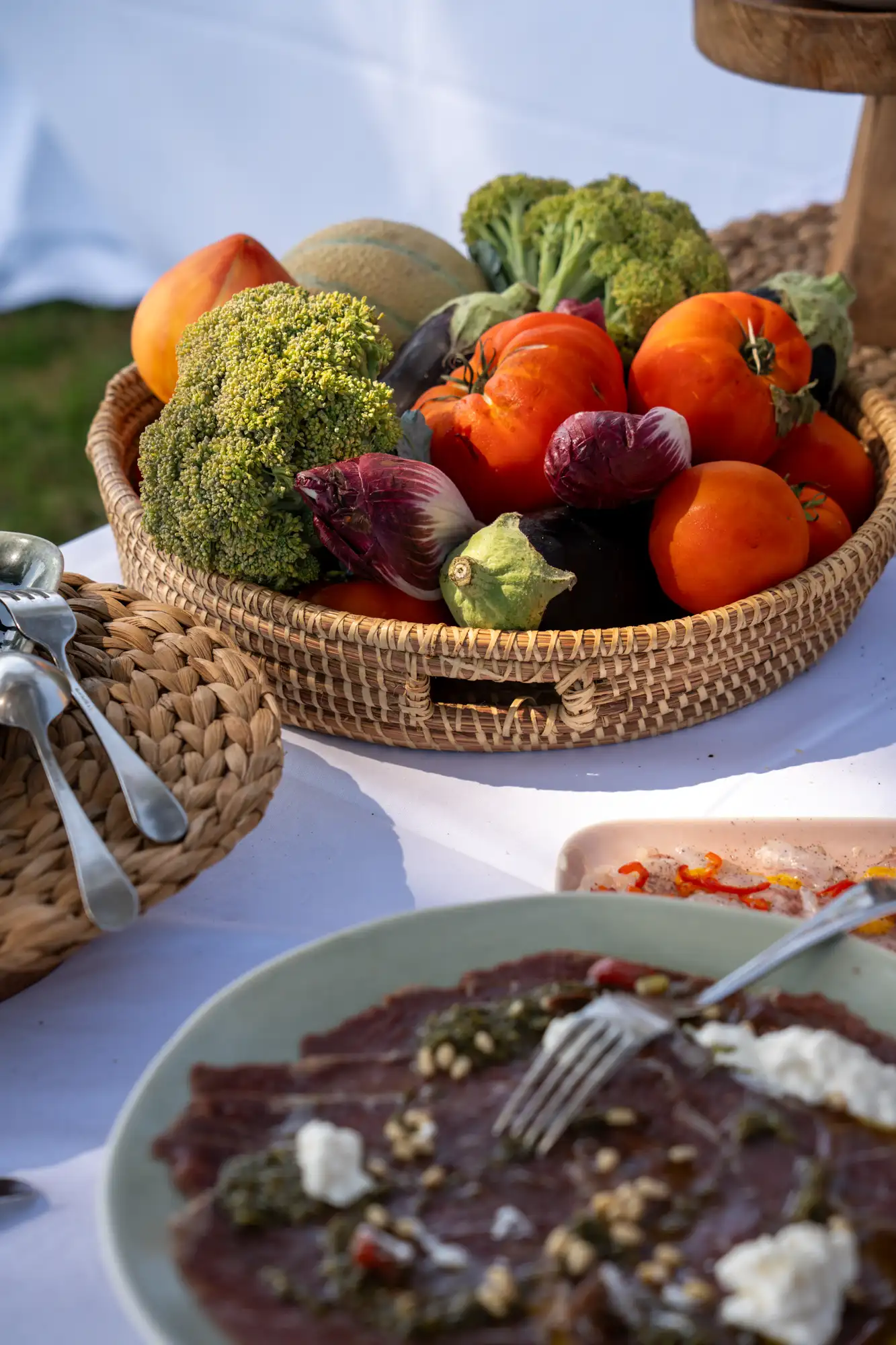Panier en osier rempli de légumes frais comprenant brocoli, tomates, aubergines, radicchio, melon et papaye, sur une table blanche à l'extérieur avec une assiette de carpaccio garnie en avant-plan.