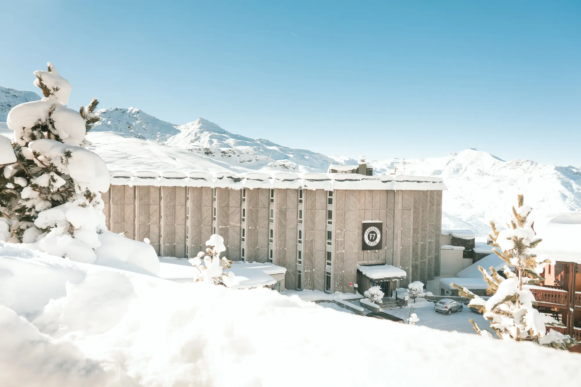 Bâtiment moderne en bois avec un panneau affichant 'F7' entouré par un paysage enneigé avec des arbres couverts de neige et des montagnes en arrière-plan sous un ciel bleu clair.