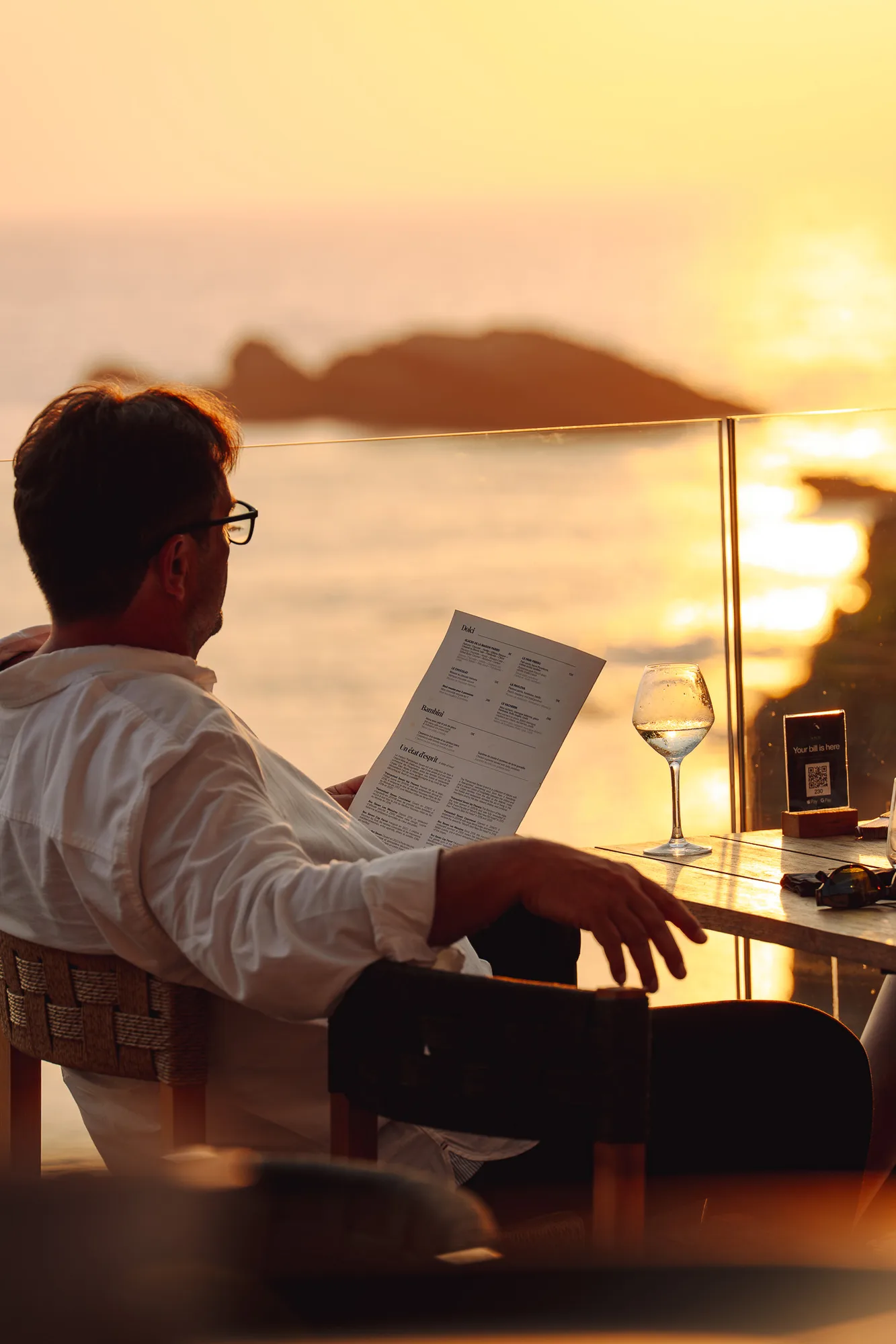 Un homme portant une chemise blanche et des lunettes est assis à une table en plein air au bord de l'eau au coucher du soleil, tenant un menu dans une main, avec un verre de vin blanc sur la table à côté de lui.