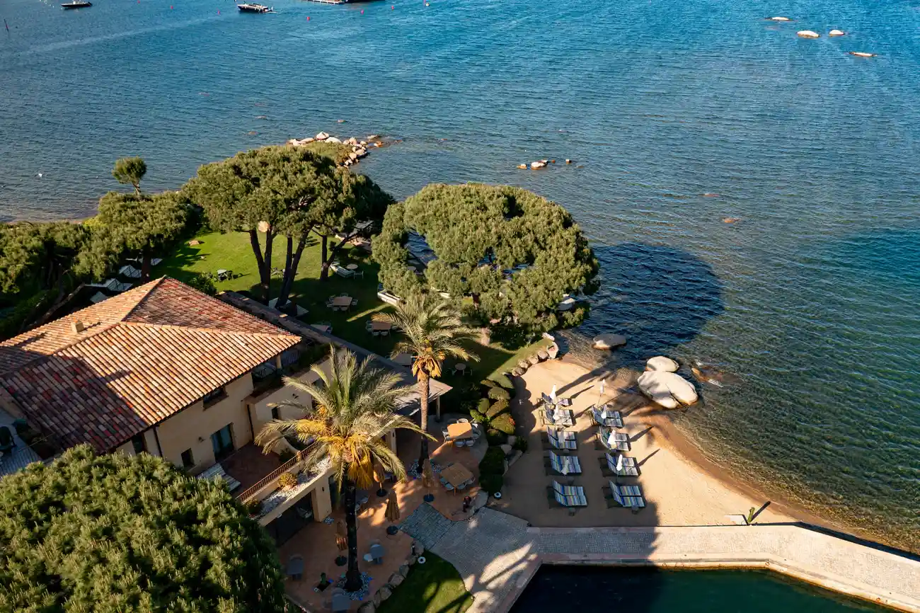 Vue aérienne d'un complexe côtier avec une maison à toit en tuiles, des palmiers et des pins, une pelouse verte avec des tables et des chaises, une plage avec plusieurs chaises longues alignées face à la mer aux eaux calmes et transparentes, ainsi que des rochers le long du rivage.