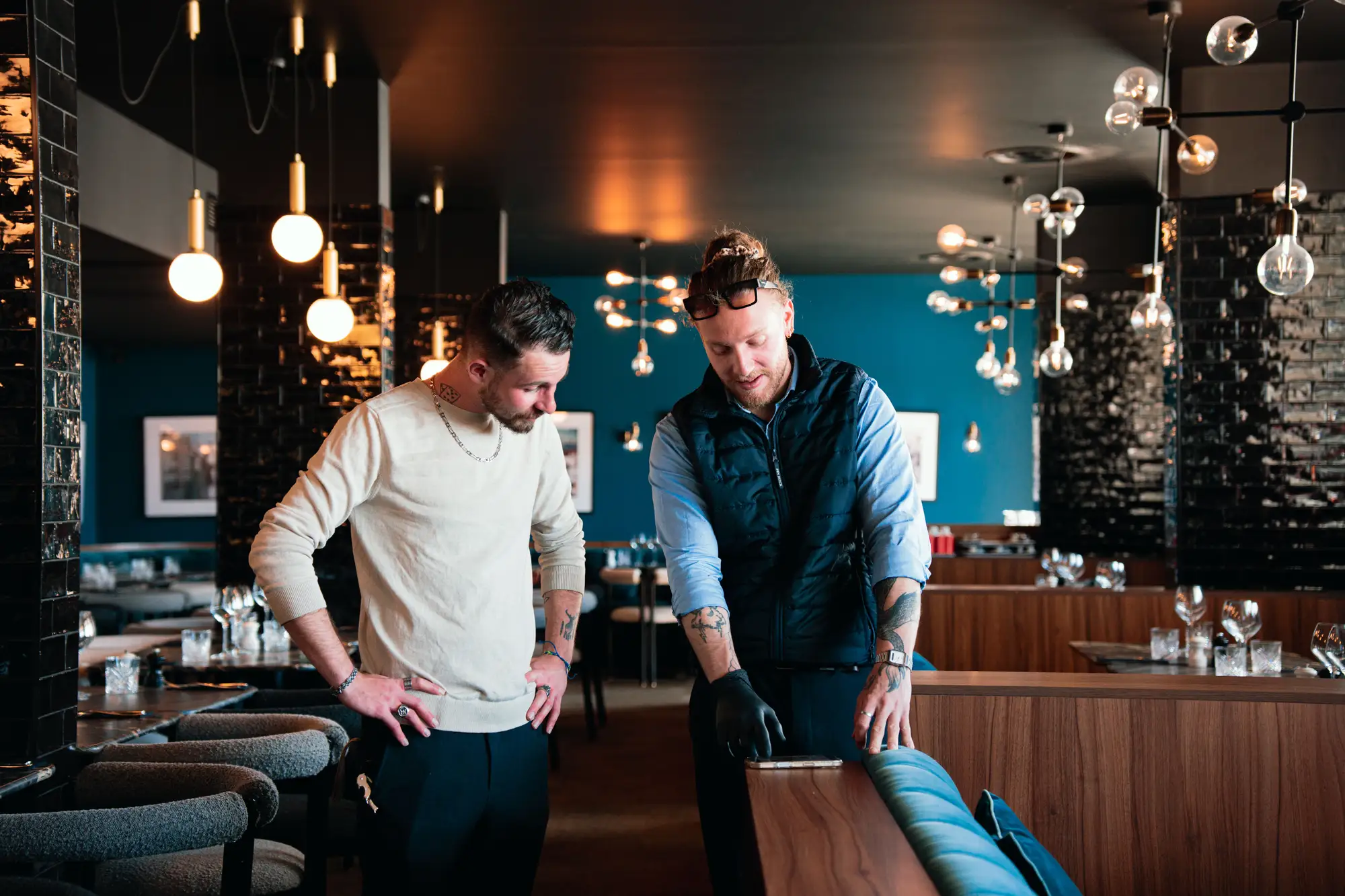 Deux hommes debout dans un restaurant sophistiqué, l'un en pull beige regardant un téléphone posé sur une banquette en bois que l'autre, portant une veste sans manches noire, montre en pointant l'écran avec un gant noir.
