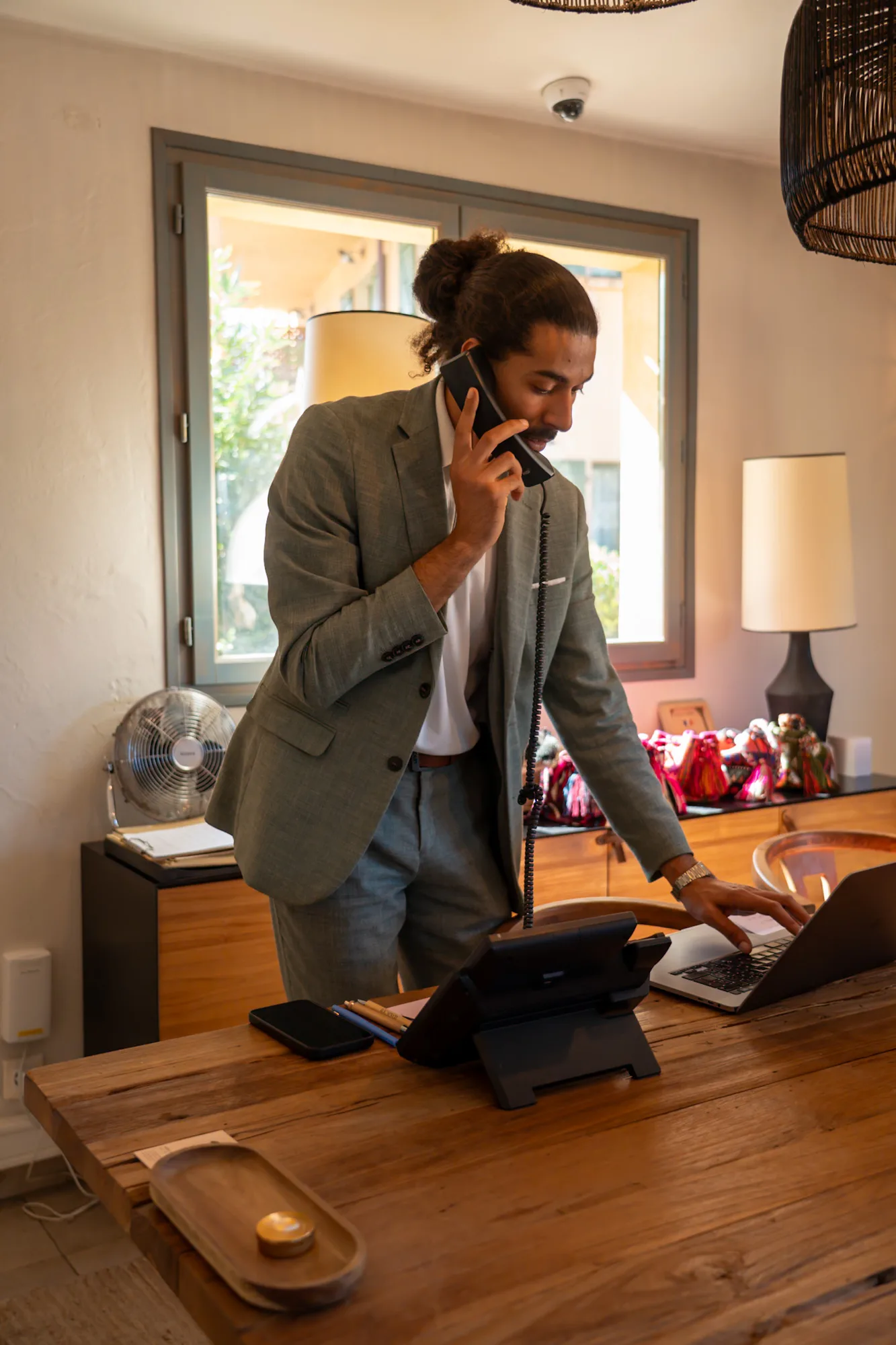 Homme d'affaires en costume gris parlant au téléphone fixe tout en utilisant un ordinateur portable dans un bureau lumineux avec une grande fenêtre, une lampe et des objets décoratifs en arrière-plan.