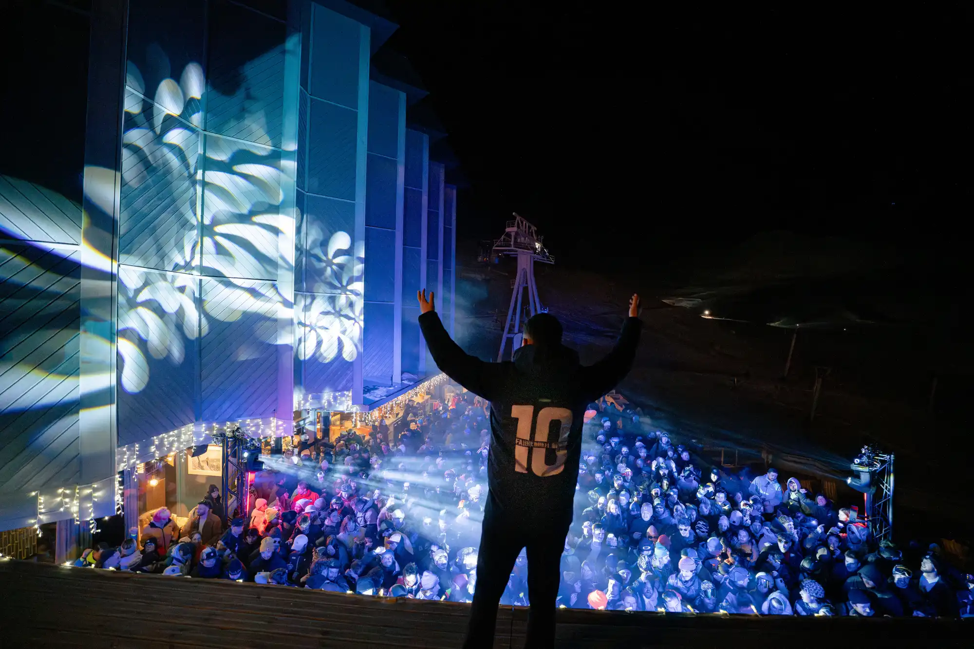 Un homme vu de dos avec les bras levés devant une foule nombreuse et éclairée lors d'une soirée extérieure nocturne avec des décorations lumineuses et des projections lumineuses sur un bâtiment adjacent.
