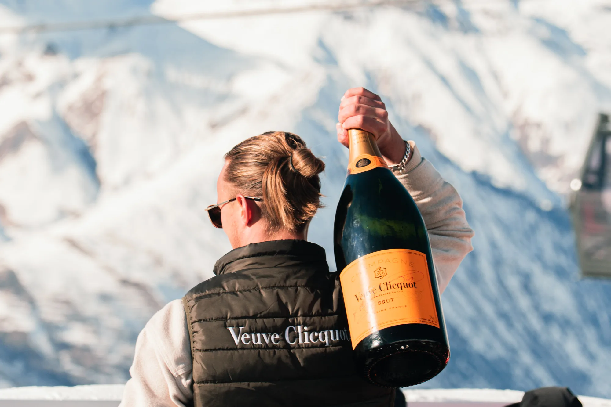 Person de dos tenant une grande bouteille de champagne Veuve Clicquot avec un paysage de montagnes enneigées en arrière-plan.