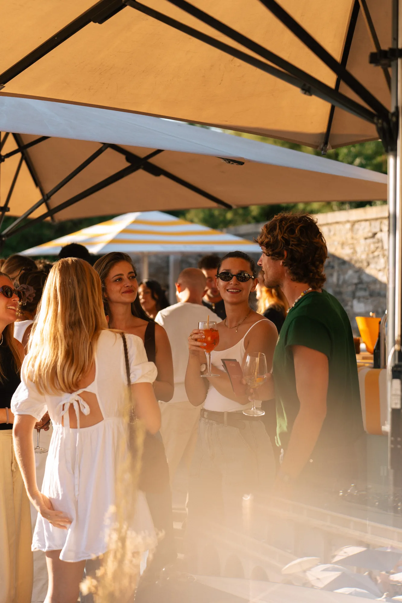 Un groupe de personnes souriantes discutant sous de grands parasols lors d'une réunion en extérieur au coucher du soleil, avec des boissons à la main.