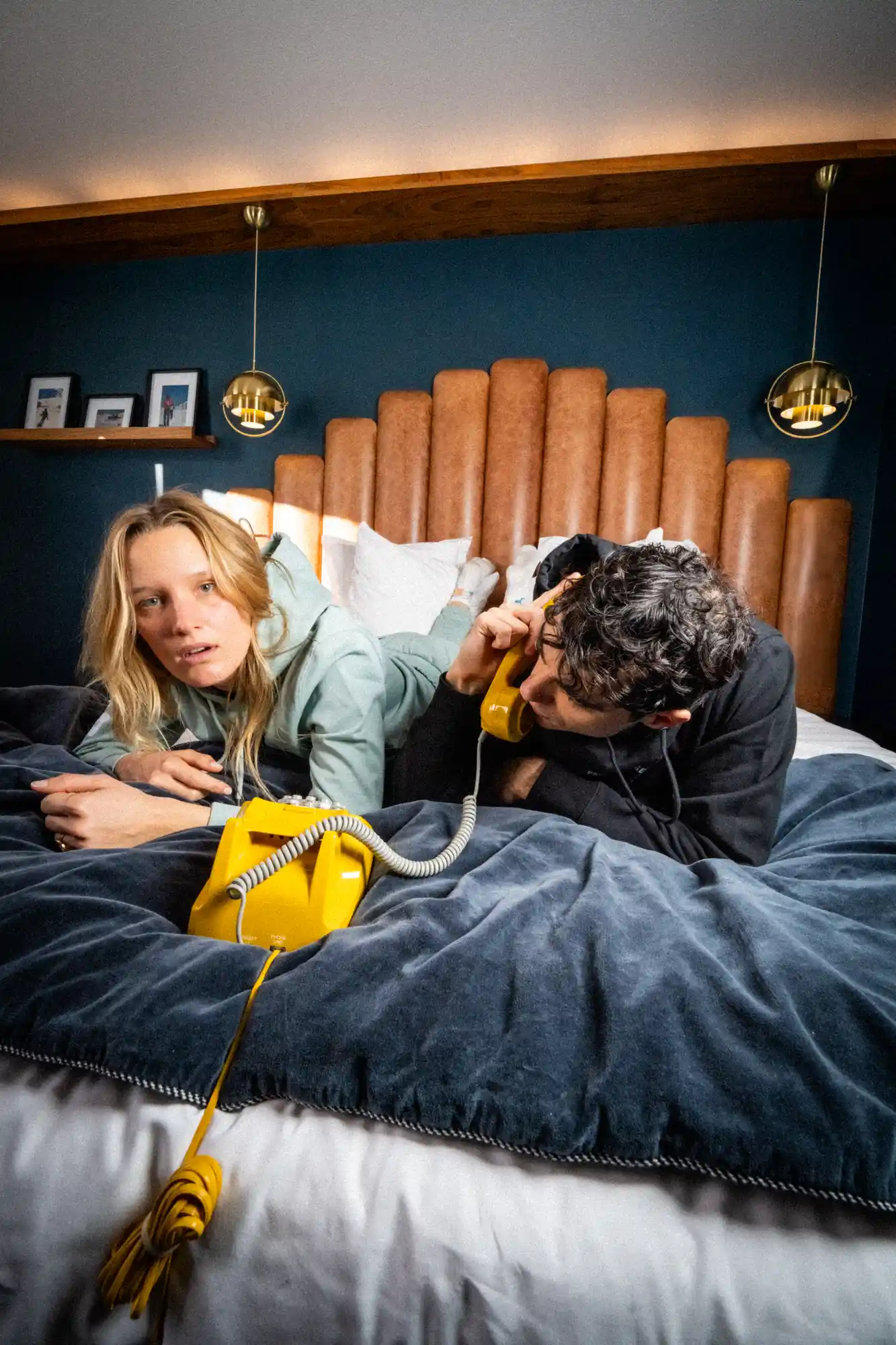 Un homme et une femme allongés sur un lit avec une tête de lit en cuir marron, le mur bleu en arrière-plan et deux lampes suspendues dorées encadrant la scène; l'homme parle dans un téléphone jaune vintage tandis que la femme regarde vers l'avant avec une expression sérieuse.