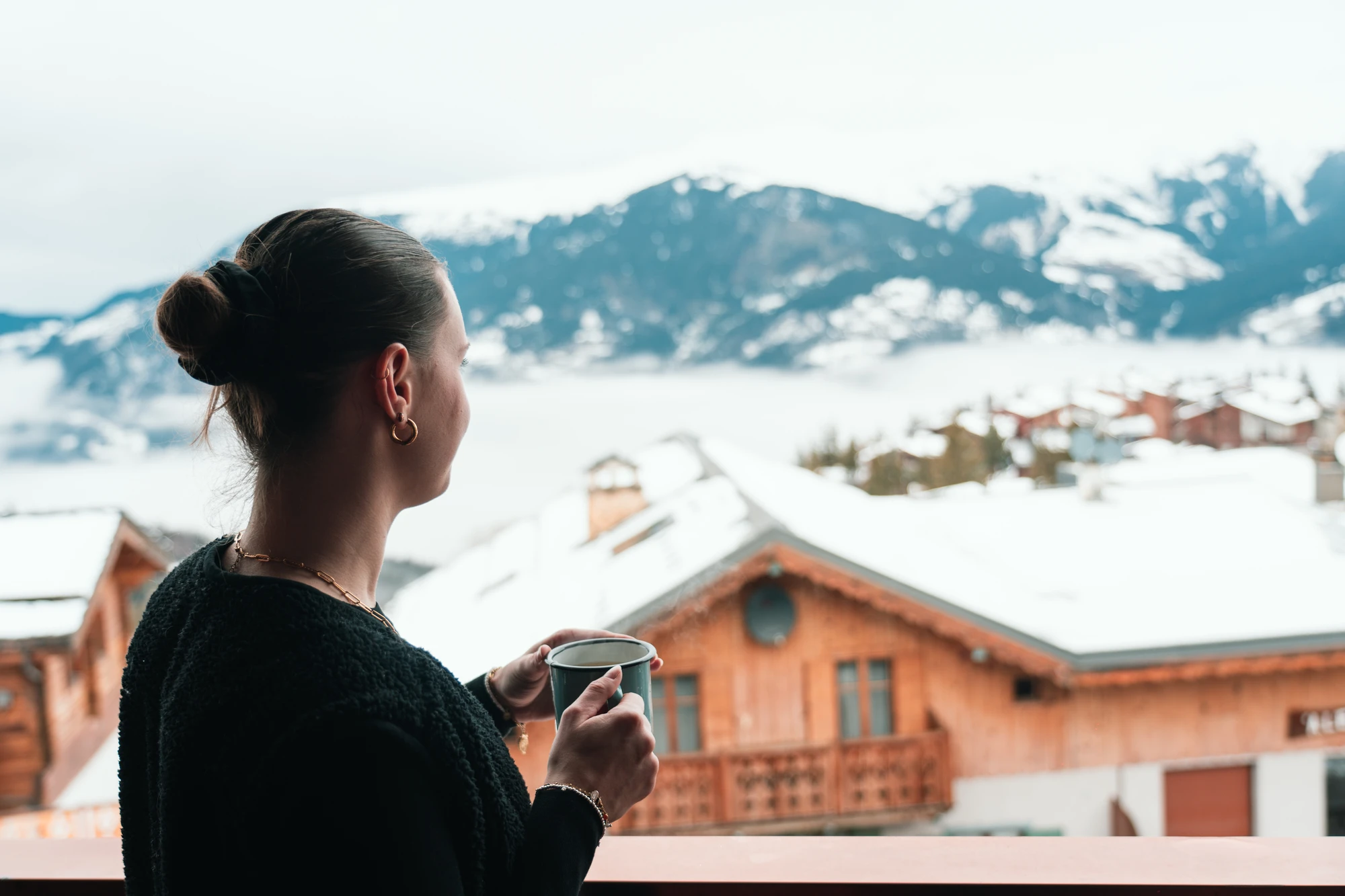 Une femme de dos tenant une tasse, regardant un paysage de montagne enneigée avec des chalets en bois au premier plan.