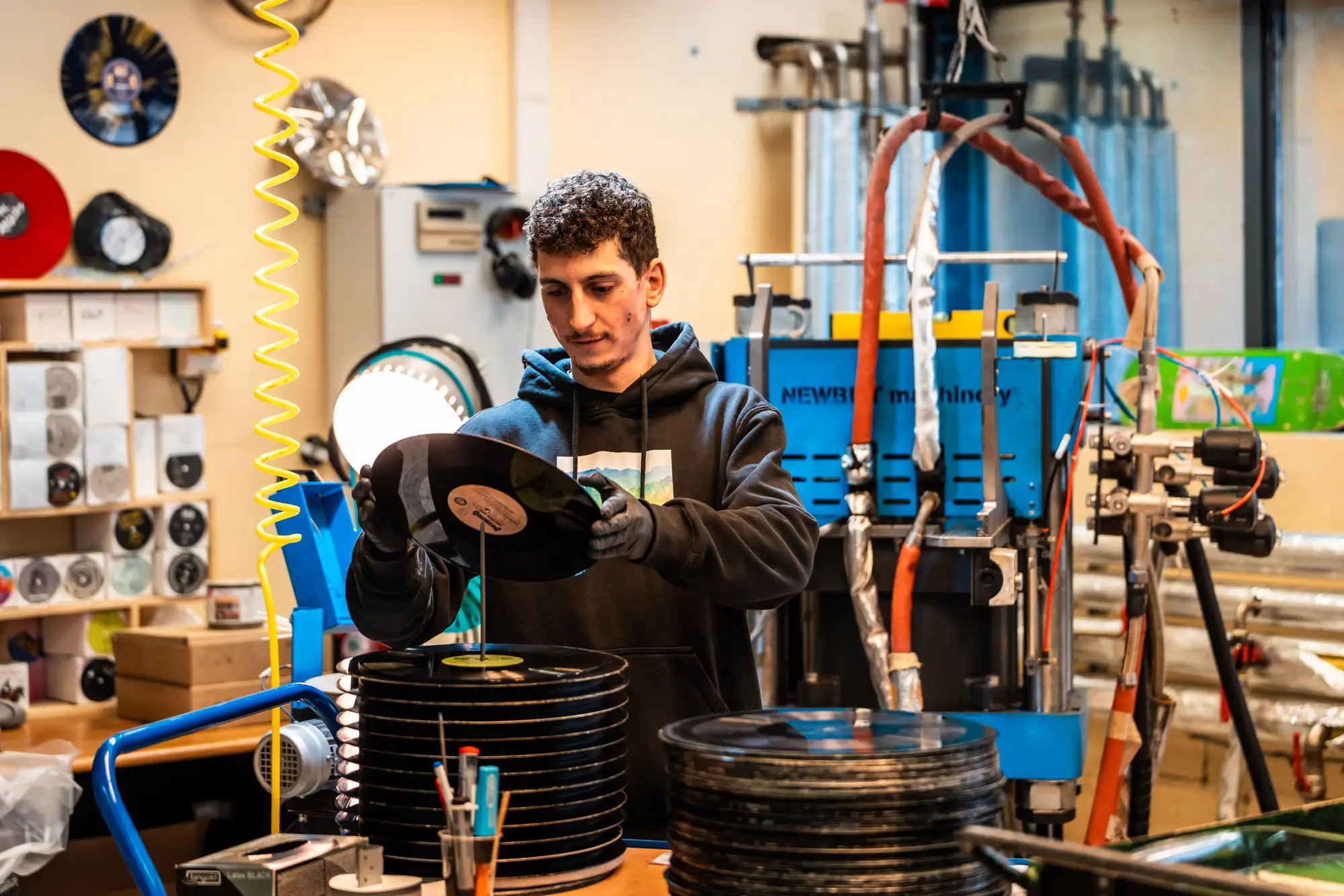 Un homme portant un sweat à capuche noir et des gants organise des disques vinyles dans un atelier avec des machines et des piles de vinyles autour de lui.