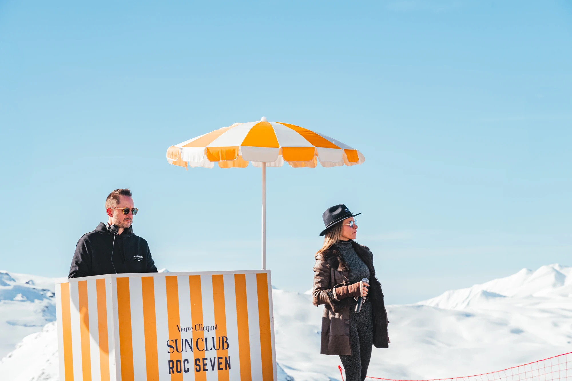Un homme portant un sweat à capuche noir et des lunettes de soleil se tient derrière un stand rayé orange et blanc avec un parasol assorti, marqué Veuve Clicquot Sun Club Roc Seven, tandis qu'une femme en manteau marron, lunettes, chapeau noir et tenant un microphone se tient à côté, avec un paysage de montagnes enneigées à l'arrière-plan.