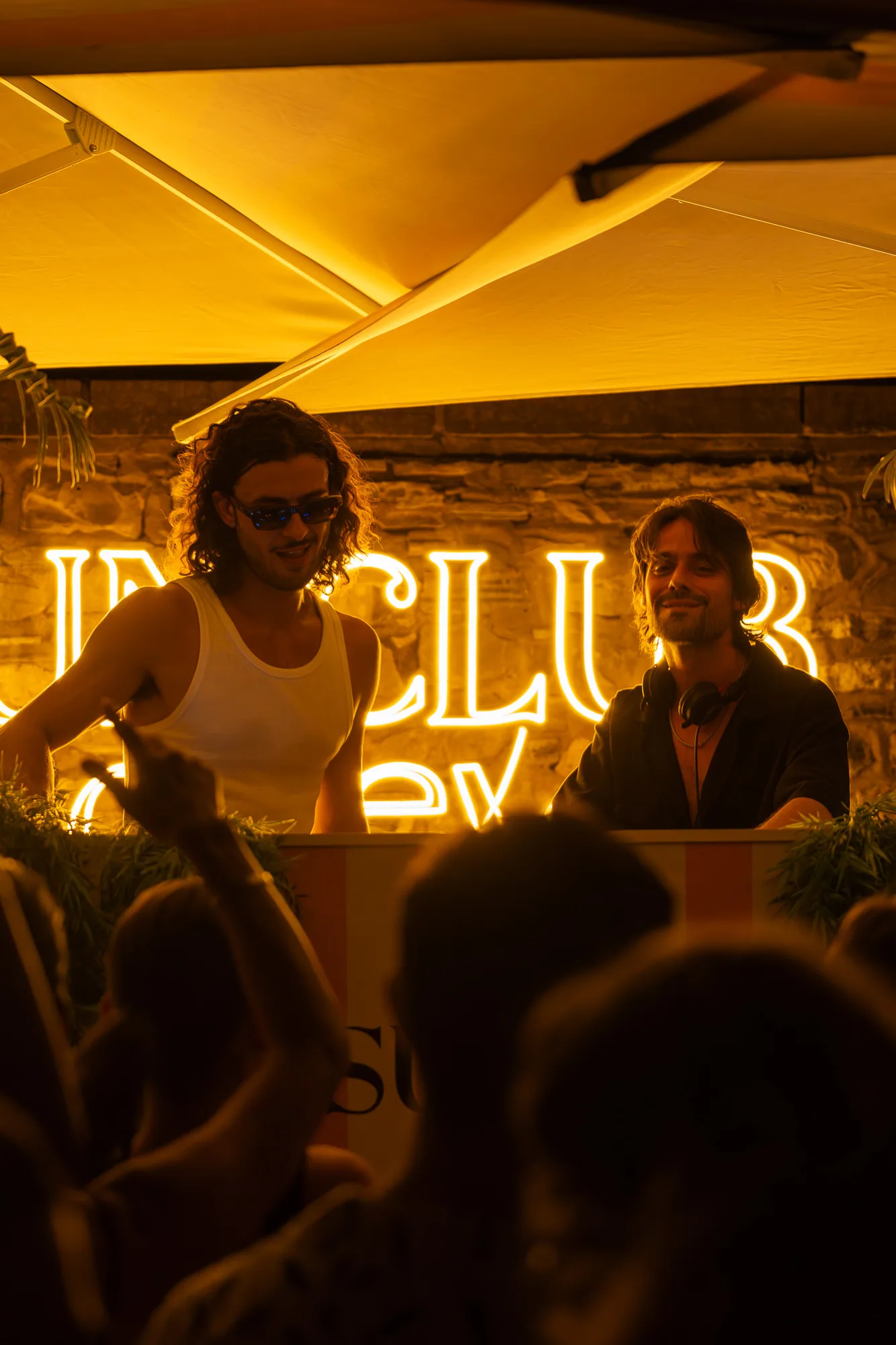 Deux DJs masculins jouent de la musique sous un auvent éclairé, avec un panneau lumineux affichant ‘‘IN CLUB‘‘ en arrière-plan, pendant que des personnes dansent au premier plan.