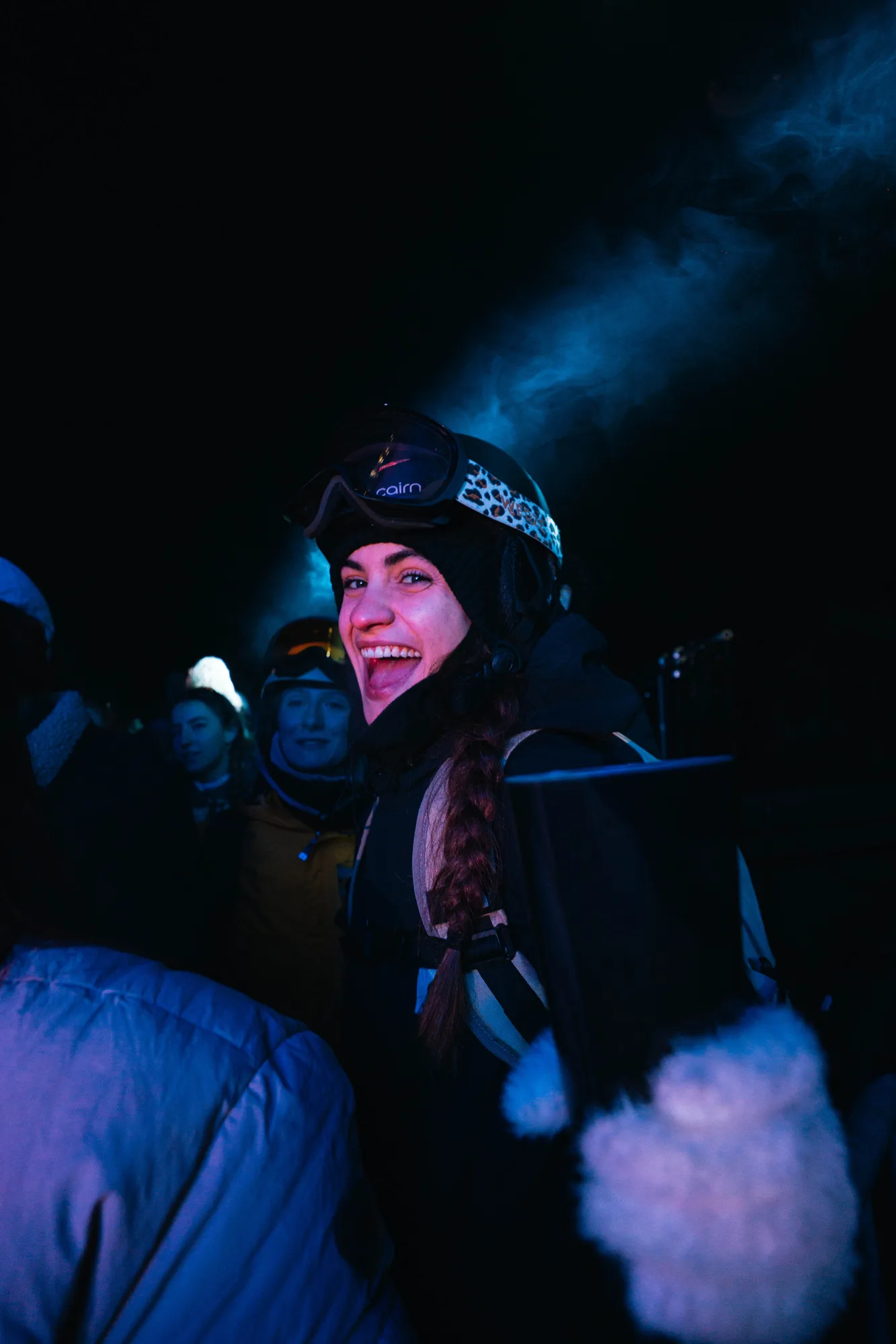 Une femme souriante portant un casque de ski noir avec des lunettes de protection et une tresse, entourée de personnes vêtues de vêtements d'hiver dans une ambiance nocturne avec une lumière bleue diffuse.