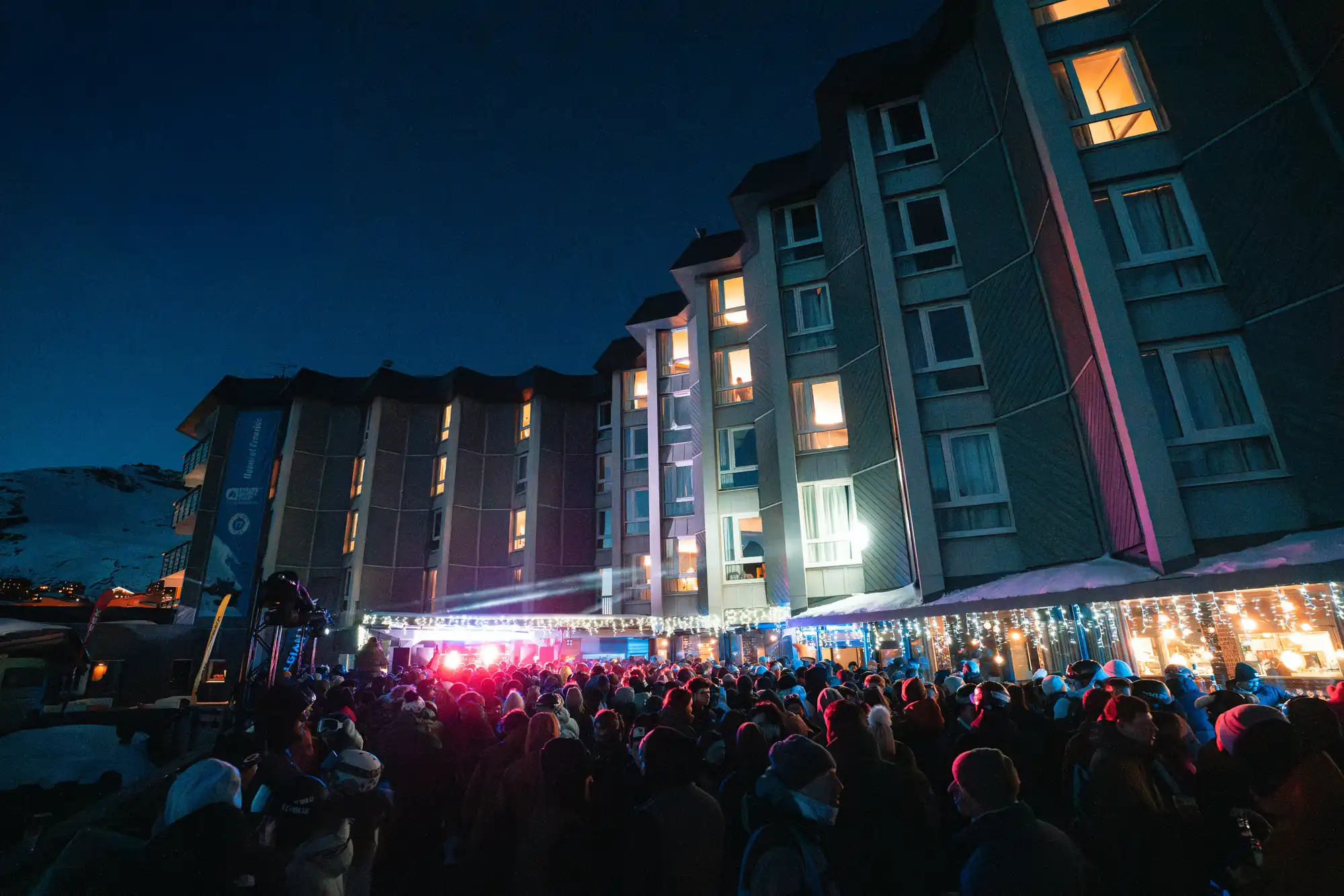 Grande foule de personnes portant des vêtements d'hiver rassemblées devant un bâtiment éclairé pendant la nuit avec des lumières décoratives scintillantes.