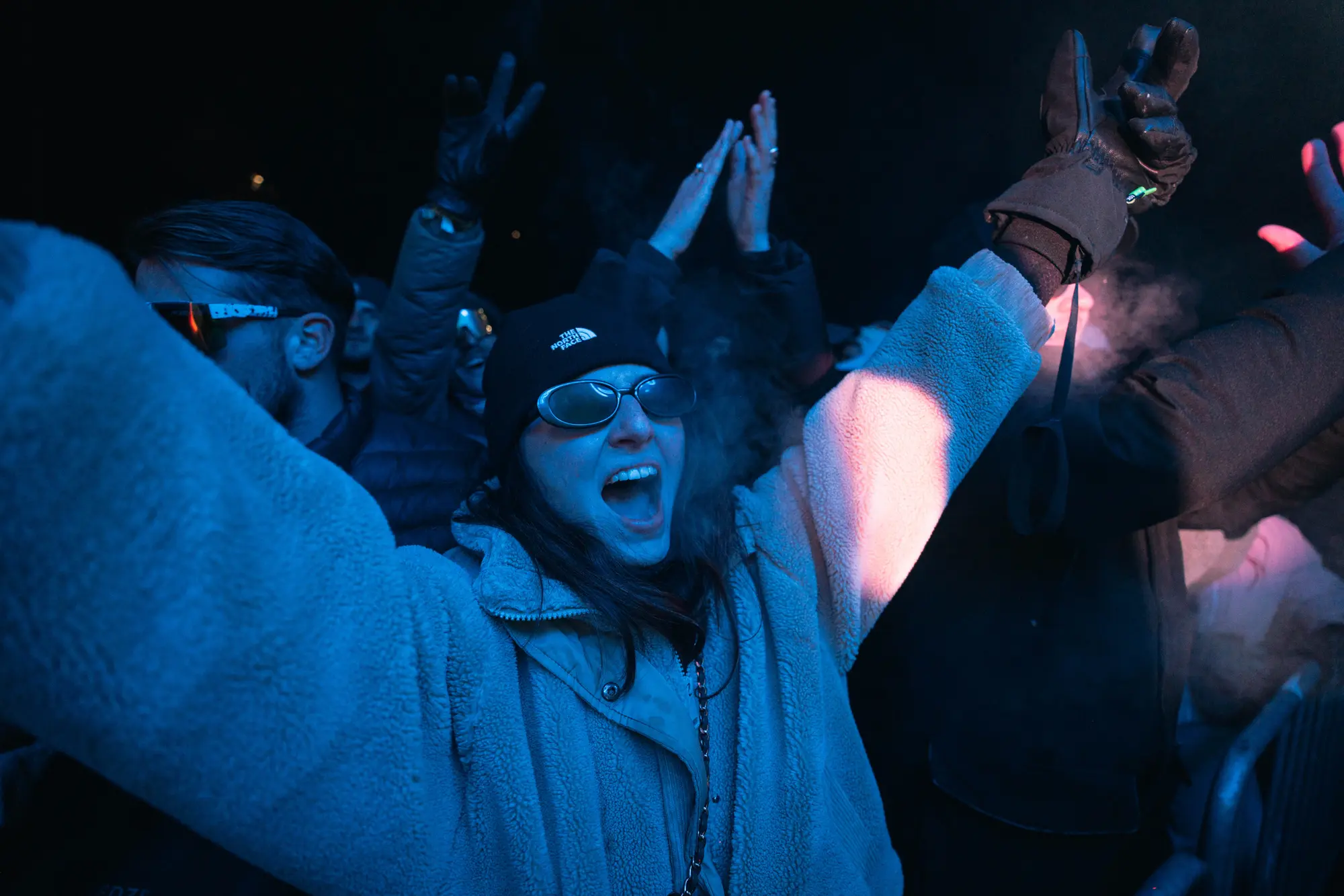 Personne portant une veste polaire épaisse, un bonnet noir The North Face et des lunettes de soleil noires, levant les bras et criant de joie dans une foule lors d'un événement nocturne avec lumière bleue.