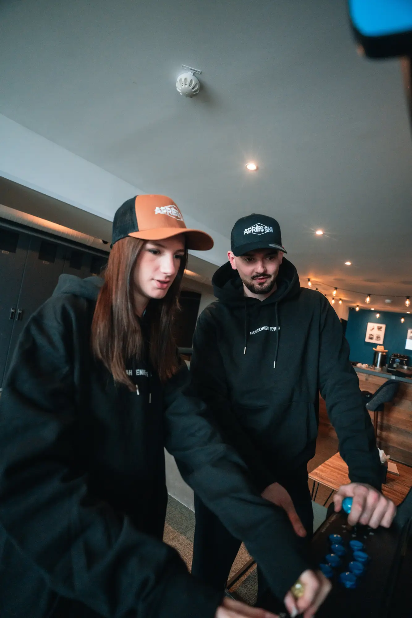 Deux personnes portant des casquettes et des sweatshirts noirs jouent à un jeu d'arcade dans une pièce intérieure éclairée par des lumières suspendues.