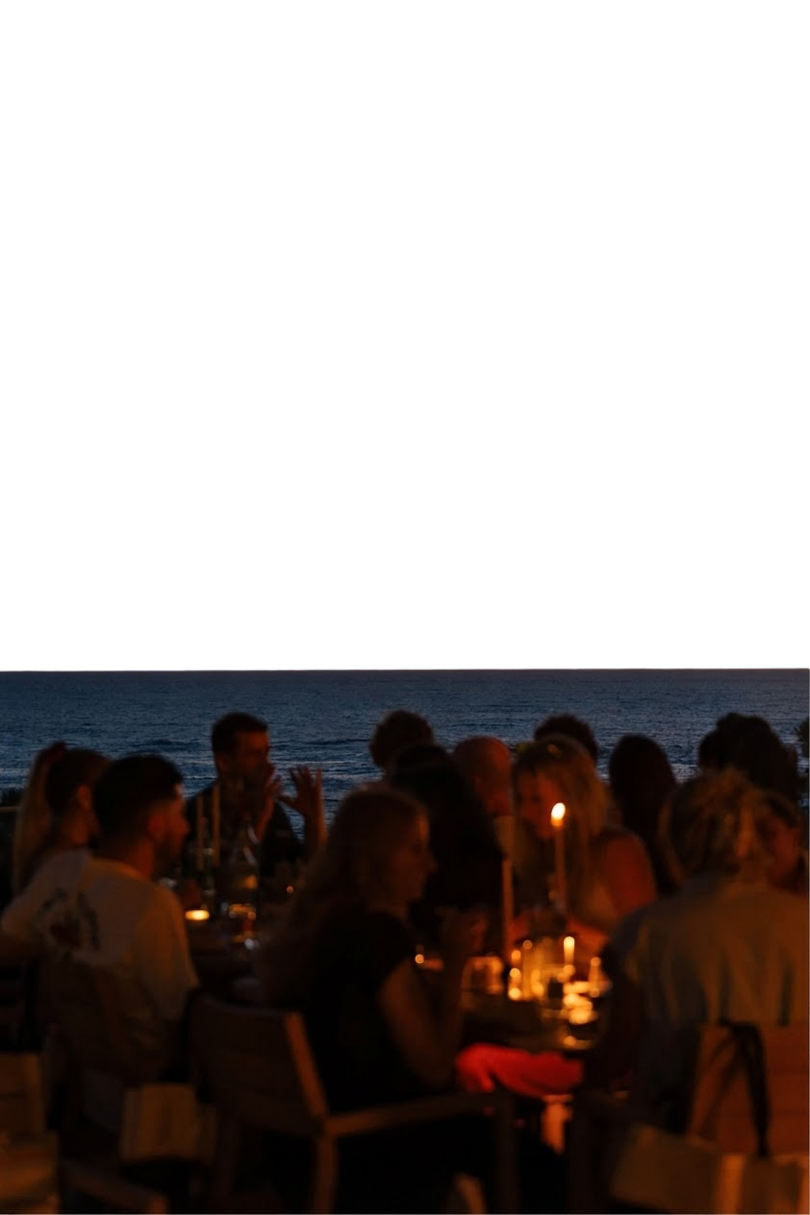 Groupe de personnes dînant ensemble au bord de la mer au crépuscule, éclairées par des bougies sur la table.