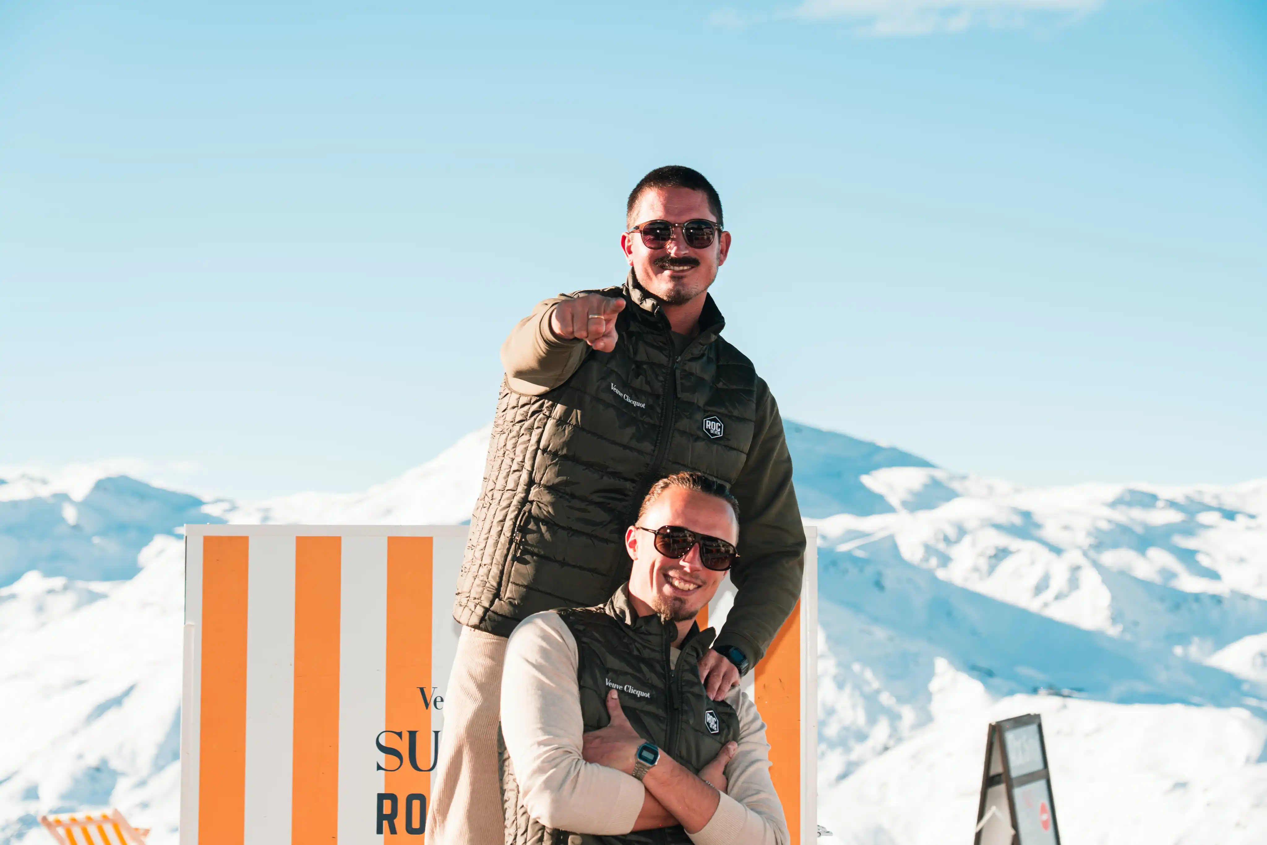 Deux hommes souriants portant des vestes sans manches noires et des lunettes de soleil posent devant un panneau rayé orange et blanc avec des montagnes enneigées en arrière-plan, sous un ciel bleu clair.