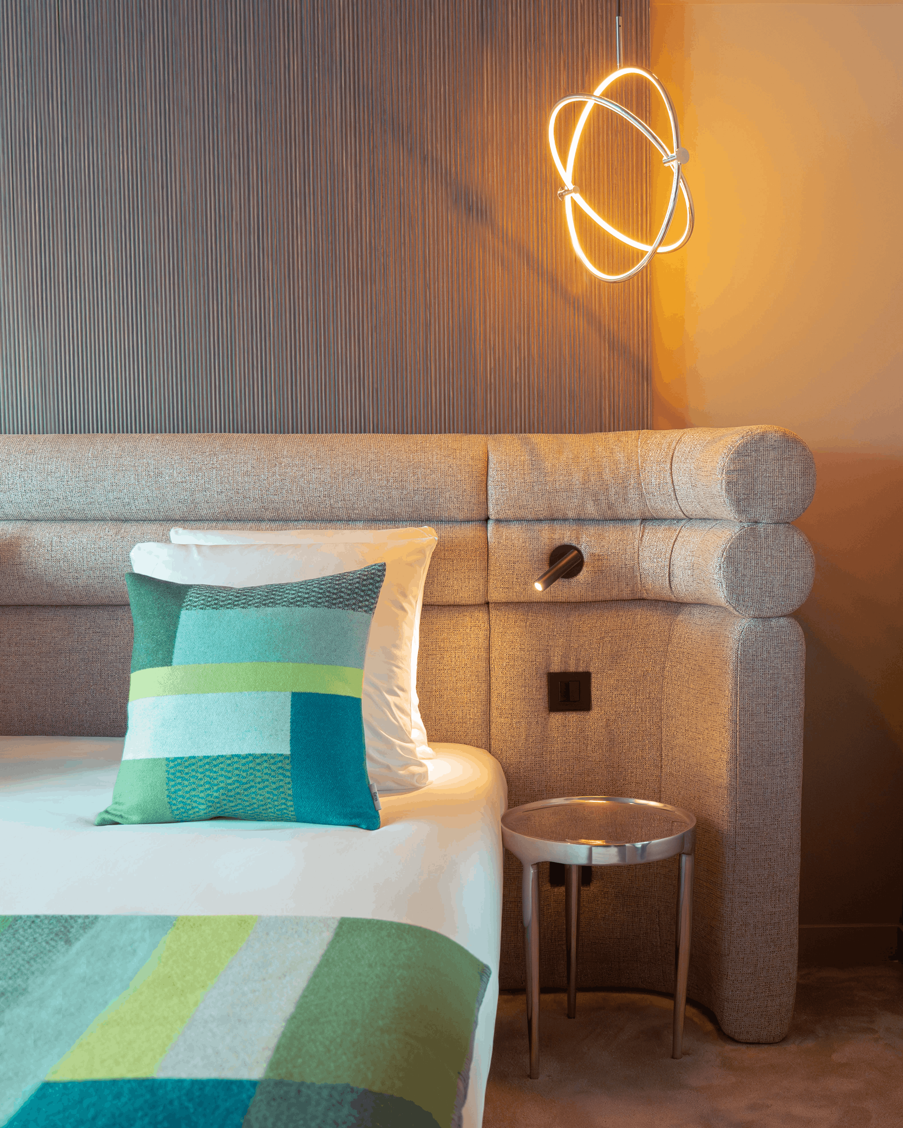 Coin d'une chambre moderne avec un lit beige, coussin décoratif vert, tête de lit grise, lampe murale design et petite table ronde en métal.