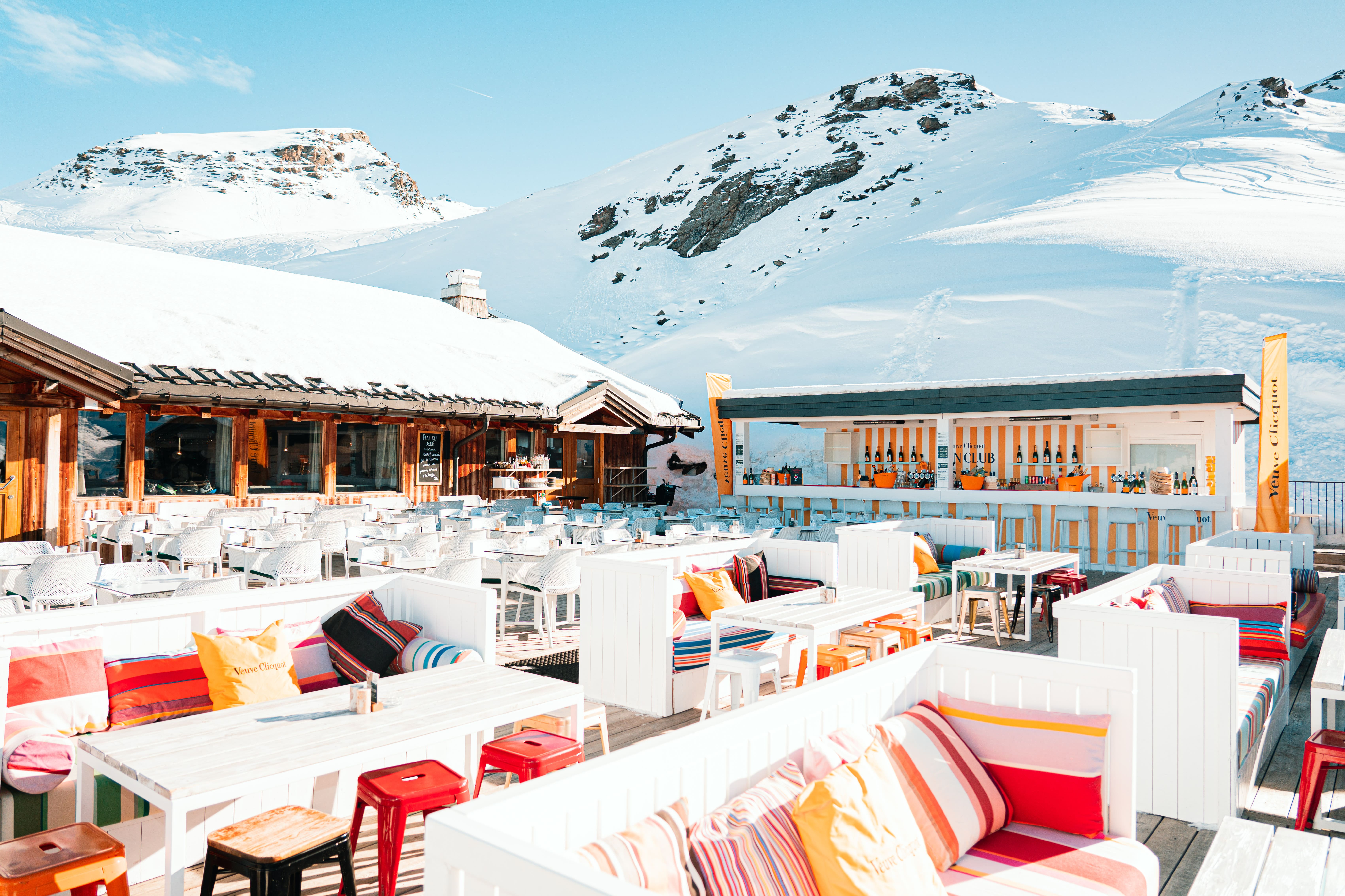 Terrasse extérieure d'un bar lounge avec des bancs blancs garnis de coussins colorés, plusieurs tables et tabourets, environnée de montagnes enneigées sous un ciel bleu clair.