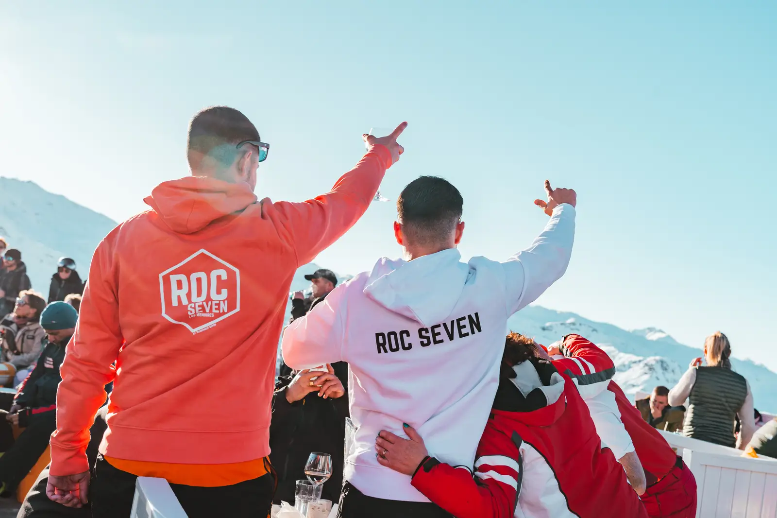 Deux hommes de dos, un portant un sweat rouge avec l'inscription RDC SEVEN, l'autre un sweat blanc avec ROC SEVEN, levant les bras en signe de célébration lors d'un rassemblement en plein air sous un ciel bleu avec des montagnes enneigées en arrière-plan.