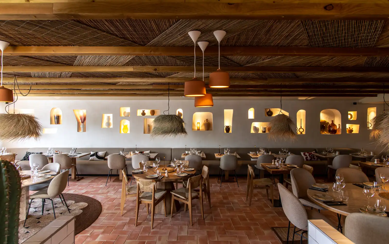 Intérieur d'un restaurant avec tables en bois, chaises confortables, des lampes suspendues en rotin et des niches éclairées dans le mur décorées de poteries et objets artisanaux, plafond avec poutres en bois et décorations naturelles.