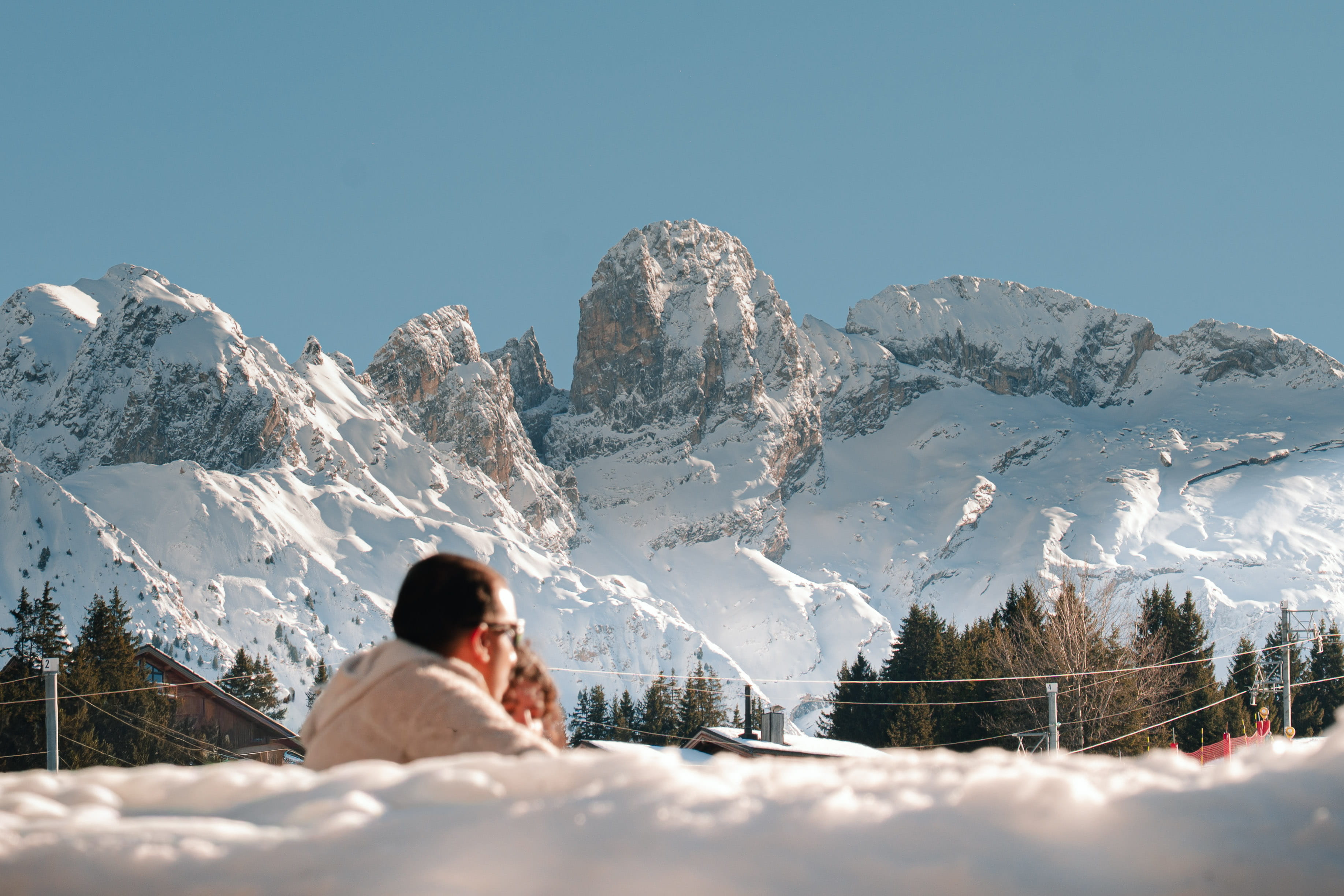Un homme portant un pull beige et des lunettes de soleil tient un enfant face à un paysage de montagnes enneigées sous un ciel bleu clair avec des arbres et des toits de maisons visibles.