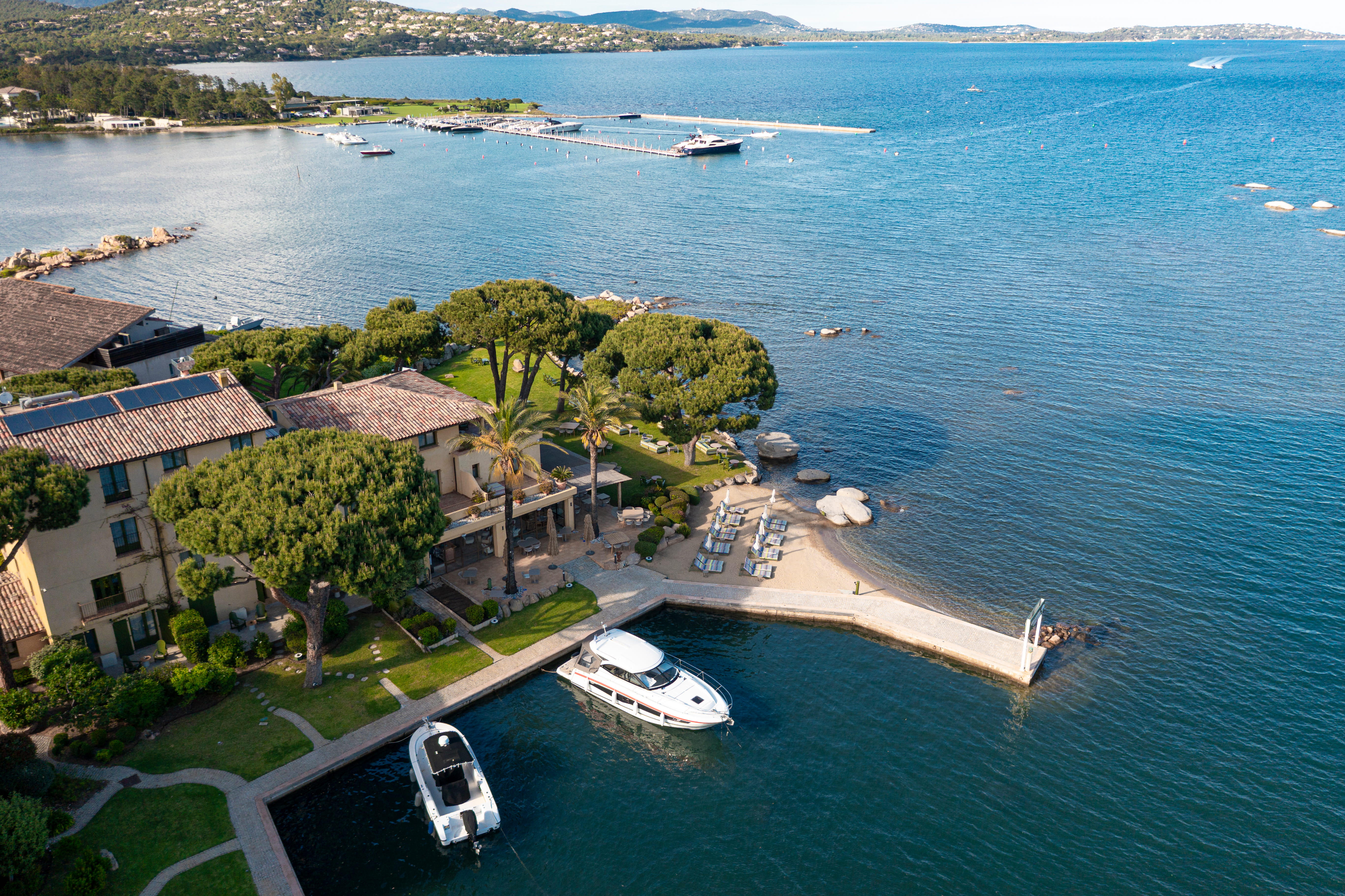 Vue aérienne d'une maison avec un toit en tuiles entourée d'arbres et d'un jardin, au bord d'une baie avec deux bateaux amarrés à un quai, une petite plage avec des chaises longues et une grande étendue d'eau bleue entourée de collines.