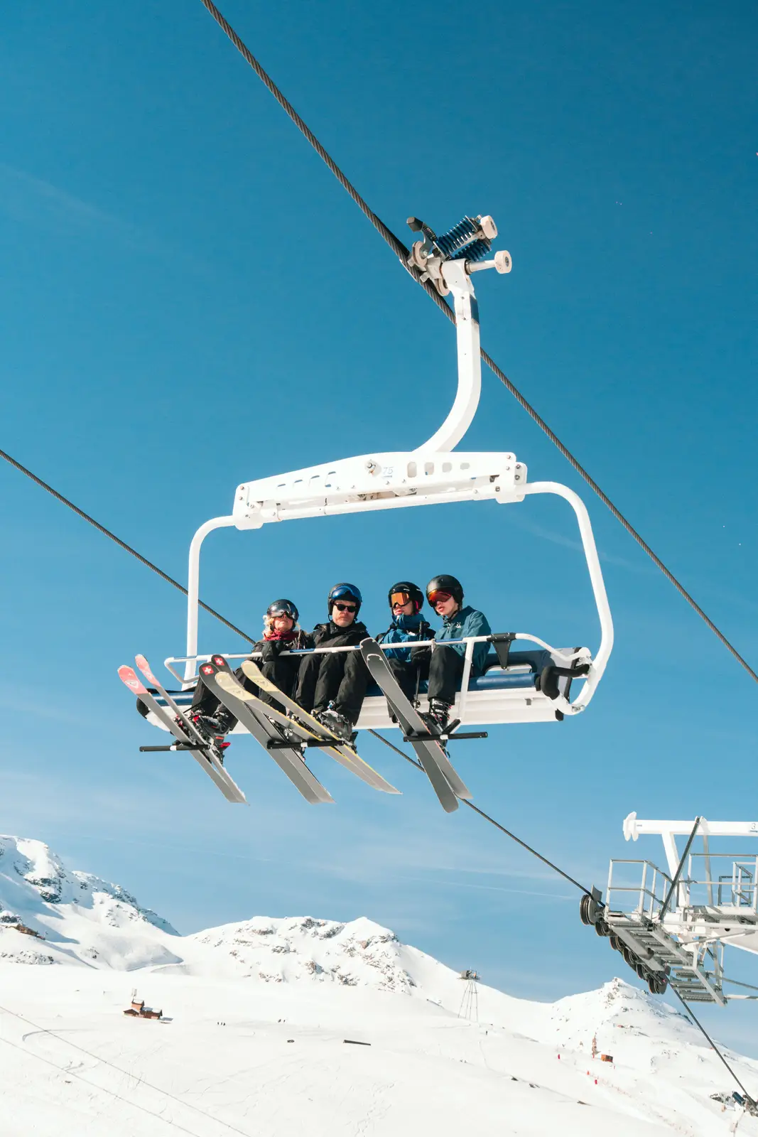 Quatre skieurs sur une télésiège blanc suspendu au-dessus d'un paysage enneigé avec des montagnes sous un ciel bleu clair.