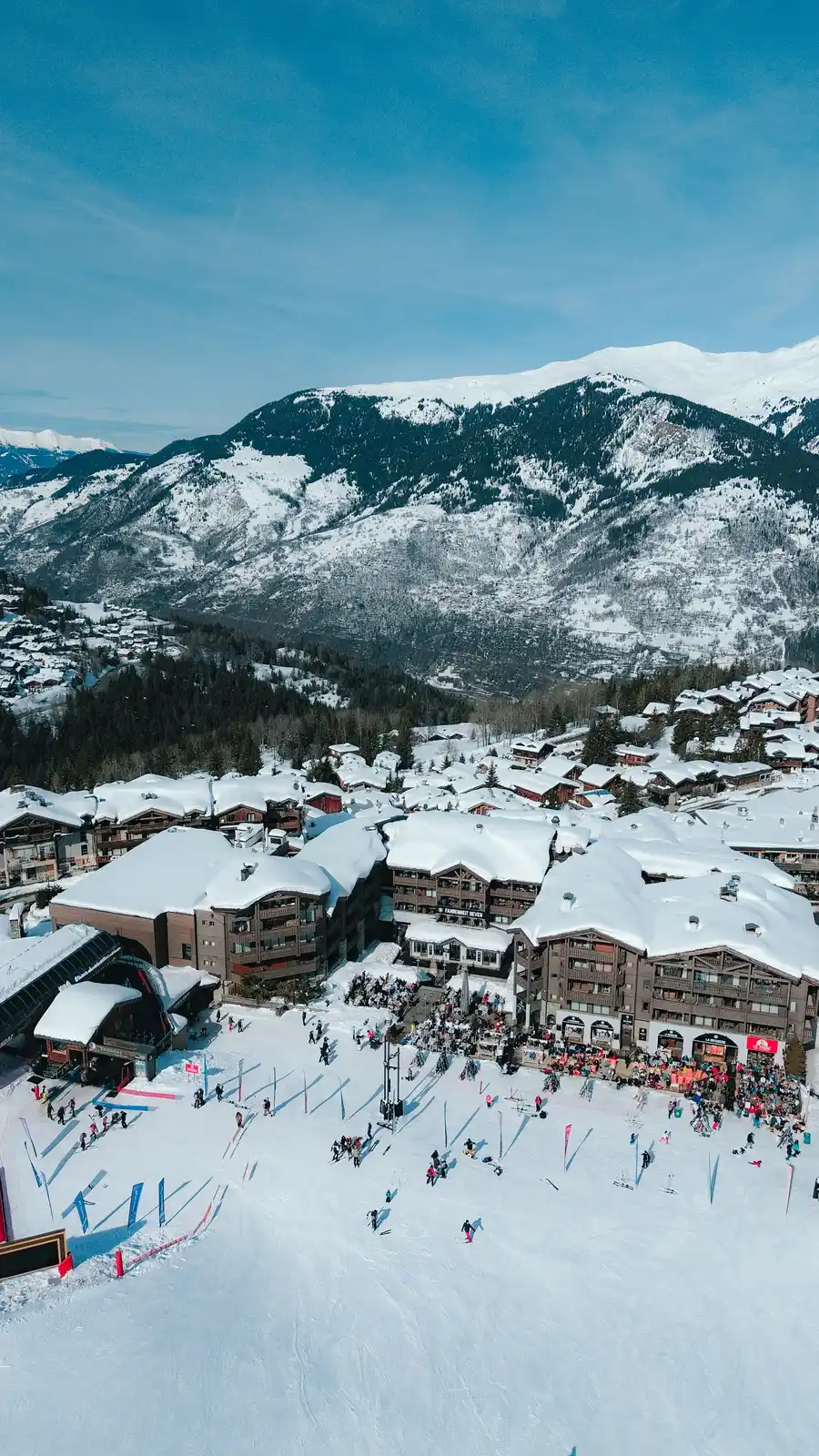 Vue aérienne d'une station de ski avec des bâtiments aux toits enneigés, des skieurs et des promeneurs sur la neige, encadrée par des montagnes enneigées sous un ciel bleu clair.