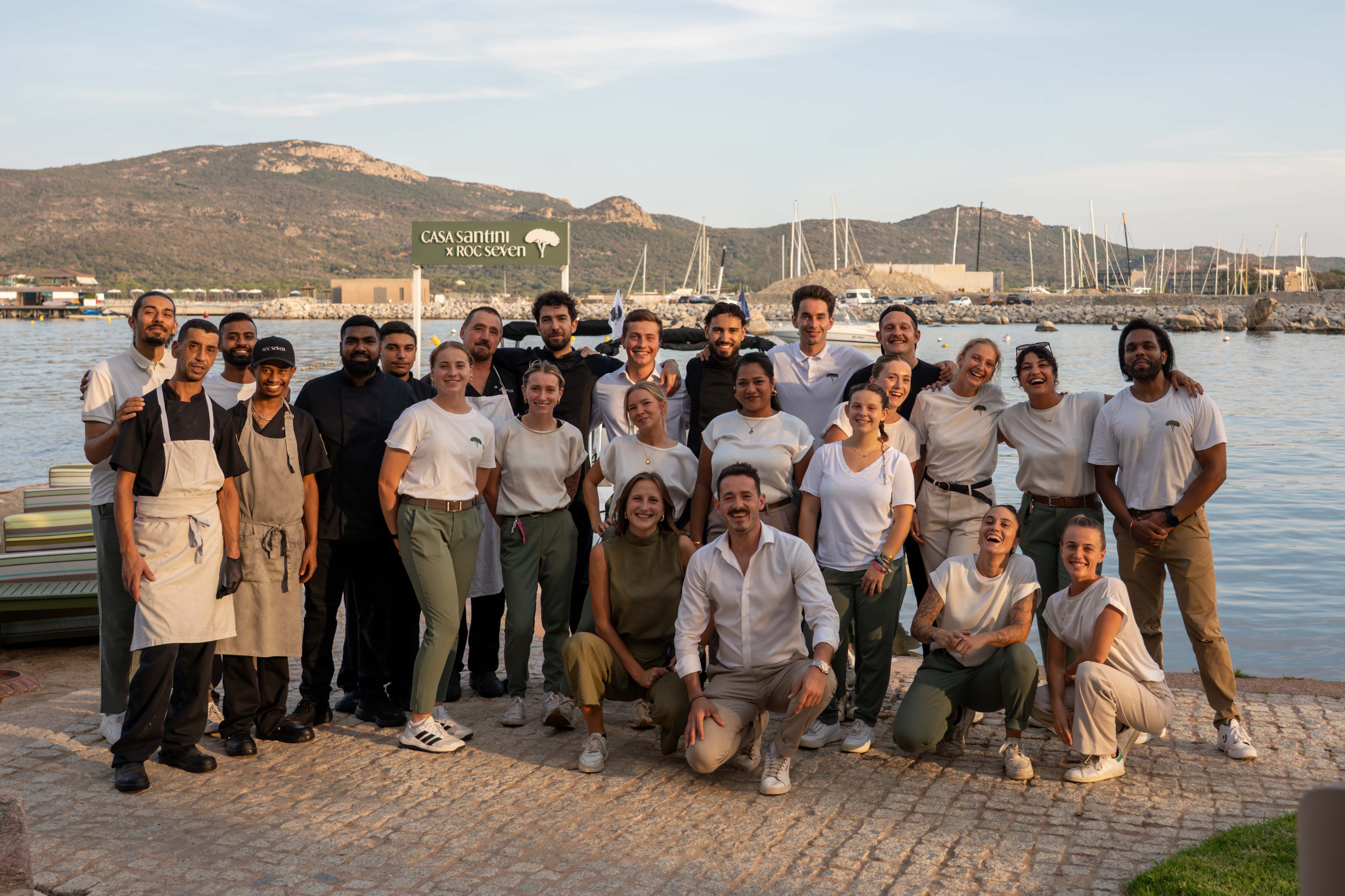 Groupe de 24 membres du personnel souriant posant devant une marina avec des bateaux et des collines en arrière-plan, certains portant des uniformes de cuisine et d'autres des tenues décontractées avec le logo Casa Santini x Roc Seven.