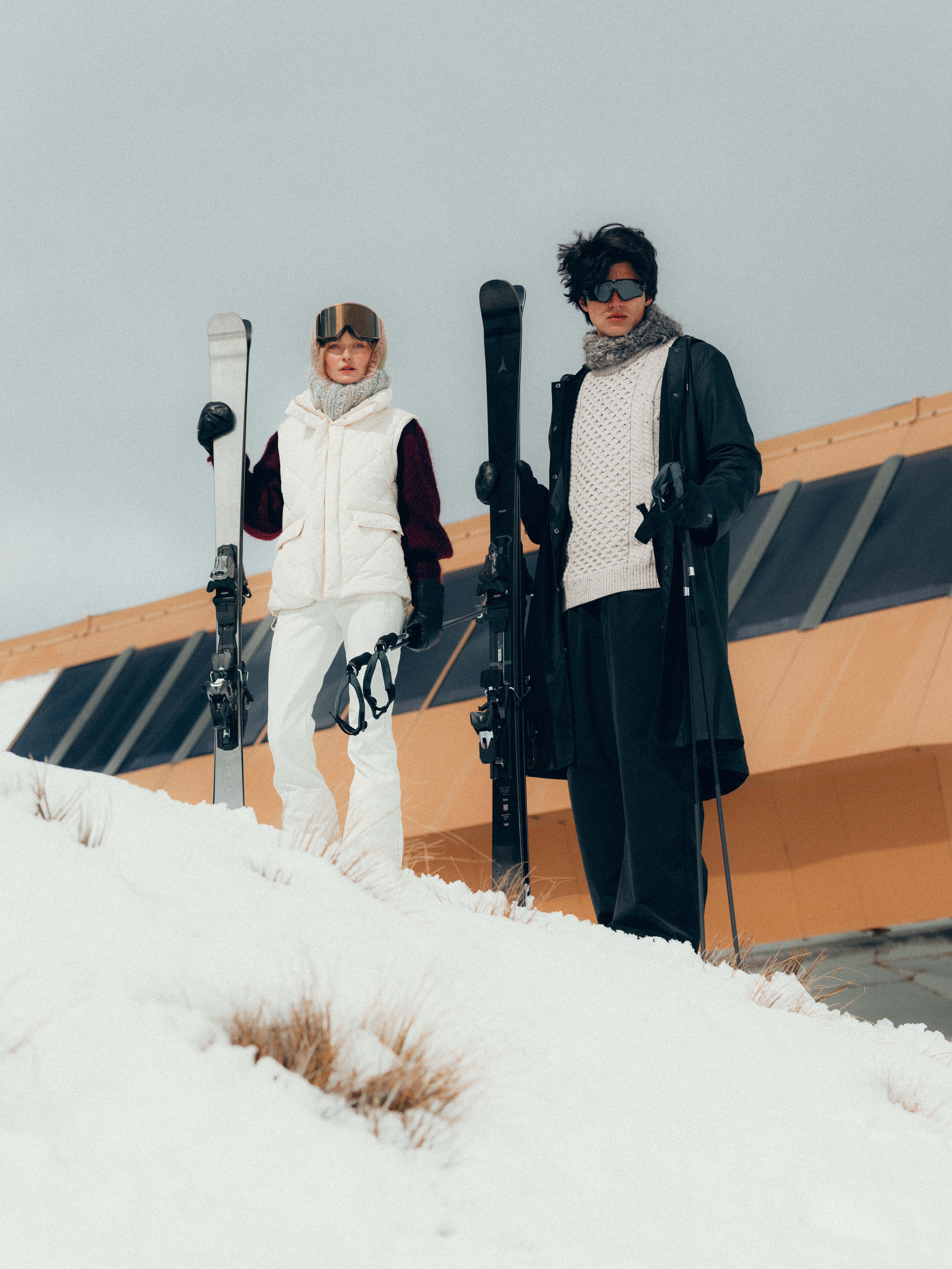 Deux skieurs, une femme à gauche en gilet blanc et pantalon de ski blanc tenant ses skis et un homme à droite en manteau noir et pull beige, portant des lunettes de ski, debout sur une pente enneigée devant un bâtiment moderne.