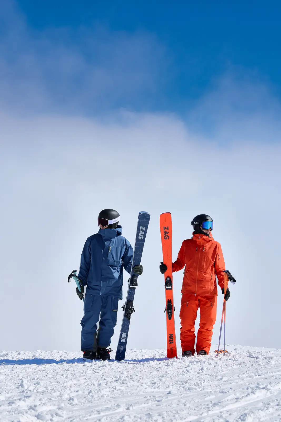 Deux skieurs debout sur la neige sous un ciel bleu, vêtus de combinaisons de ski aux couleurs bleu et orange, tenant chacun une paire de skis et des bâtons de ski.