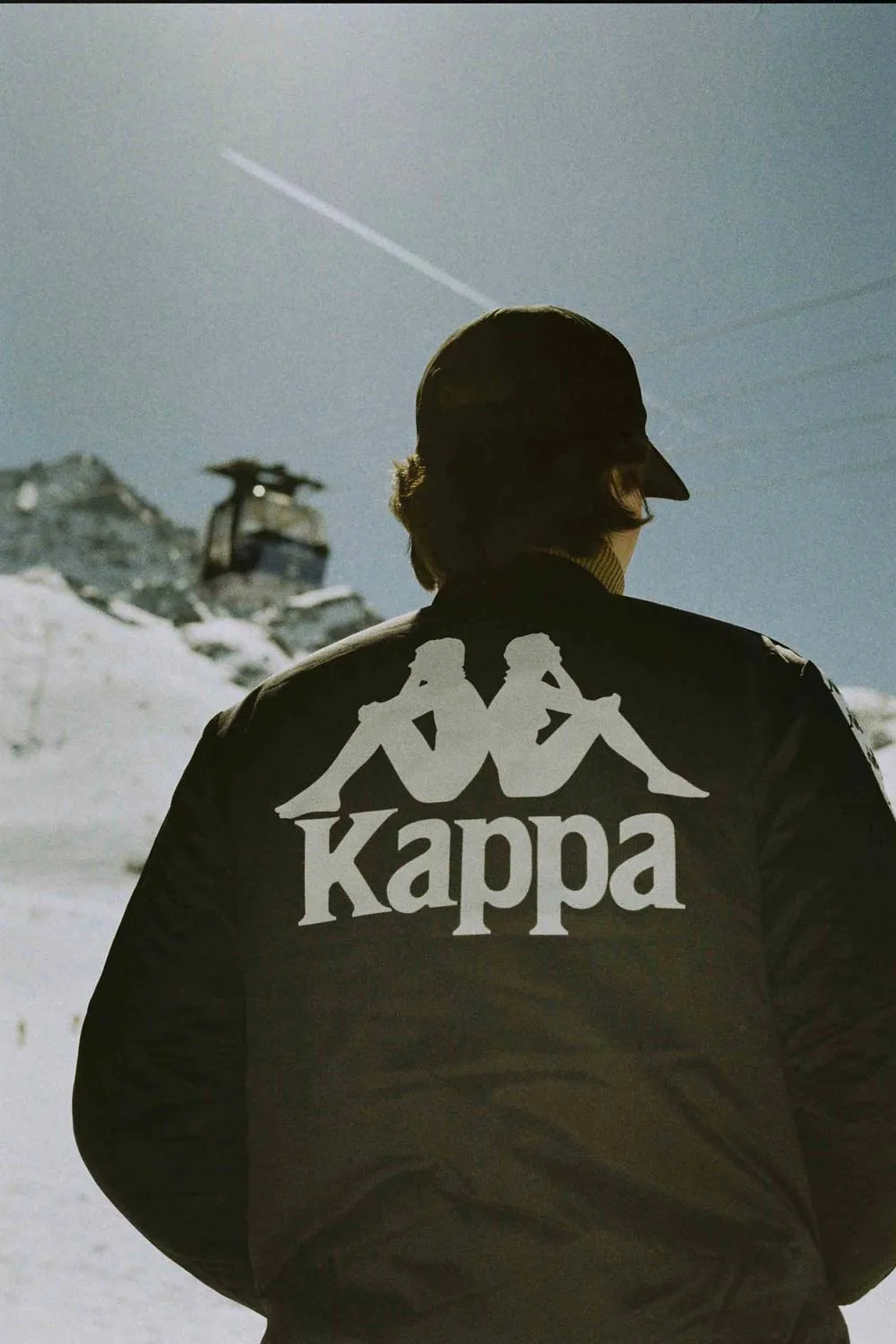 Person portant une veste noire avec le logo Kappa en blanc montrant deux silhouettes assises dos à dos, dans un paysage montagneux enneigé sous un ciel bleu clair.