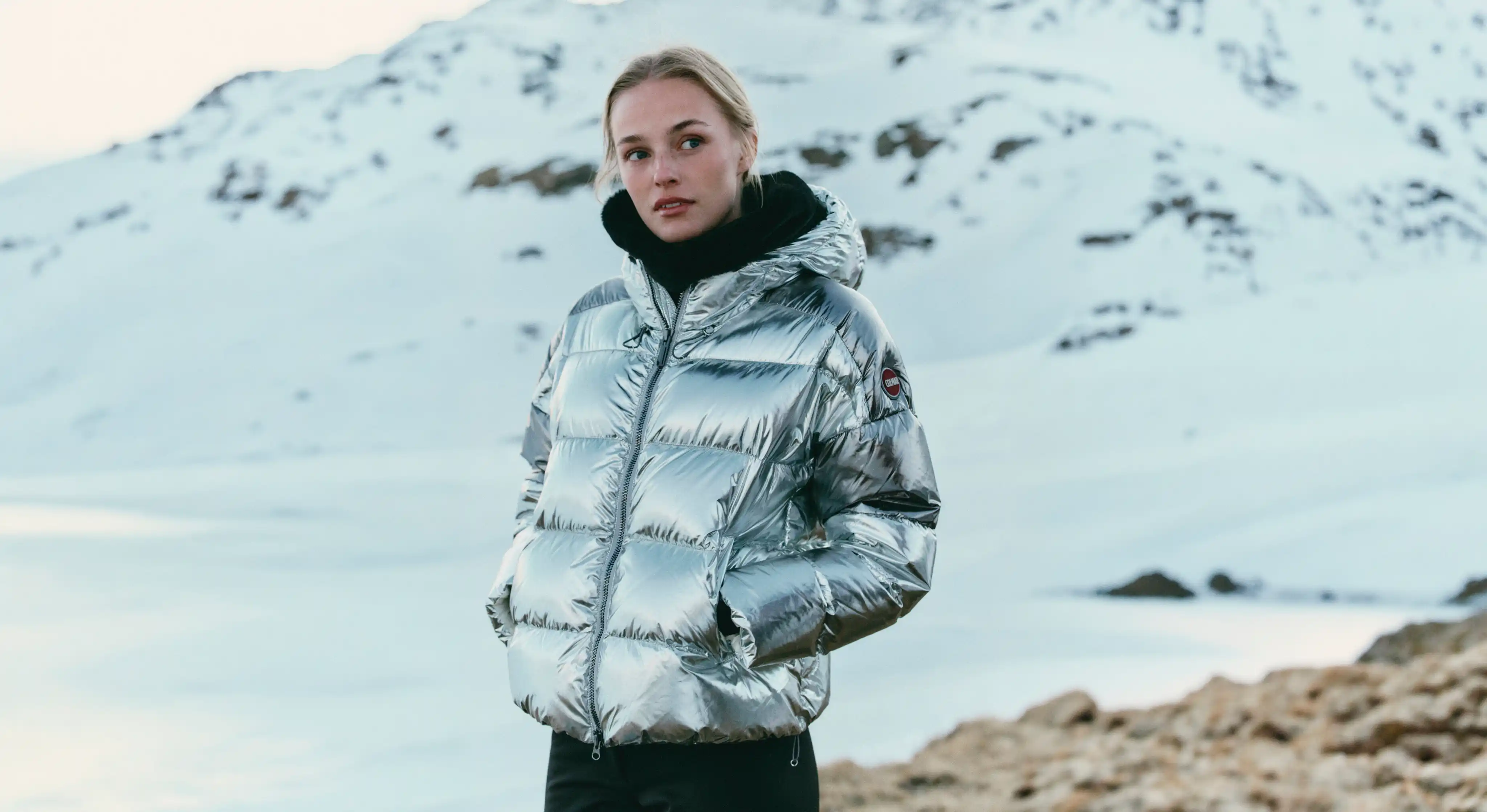 Jeune femme portant une doudoune argentée brillante et une écharpe noire, debout dans un paysage enneigé avec des montagnes en arrière-plan.