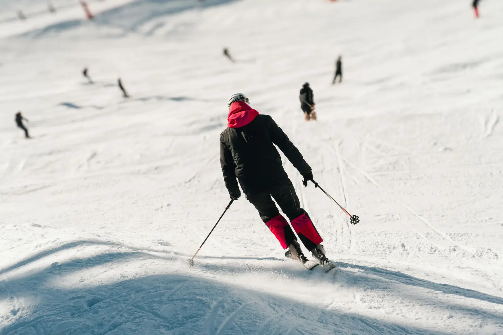 Personne vue de dos skiant en descente sur une piste enneigée, portant une veste noire avec des détails rouges et un casque, avec plusieurs autres skieurs en arrière-plan.
