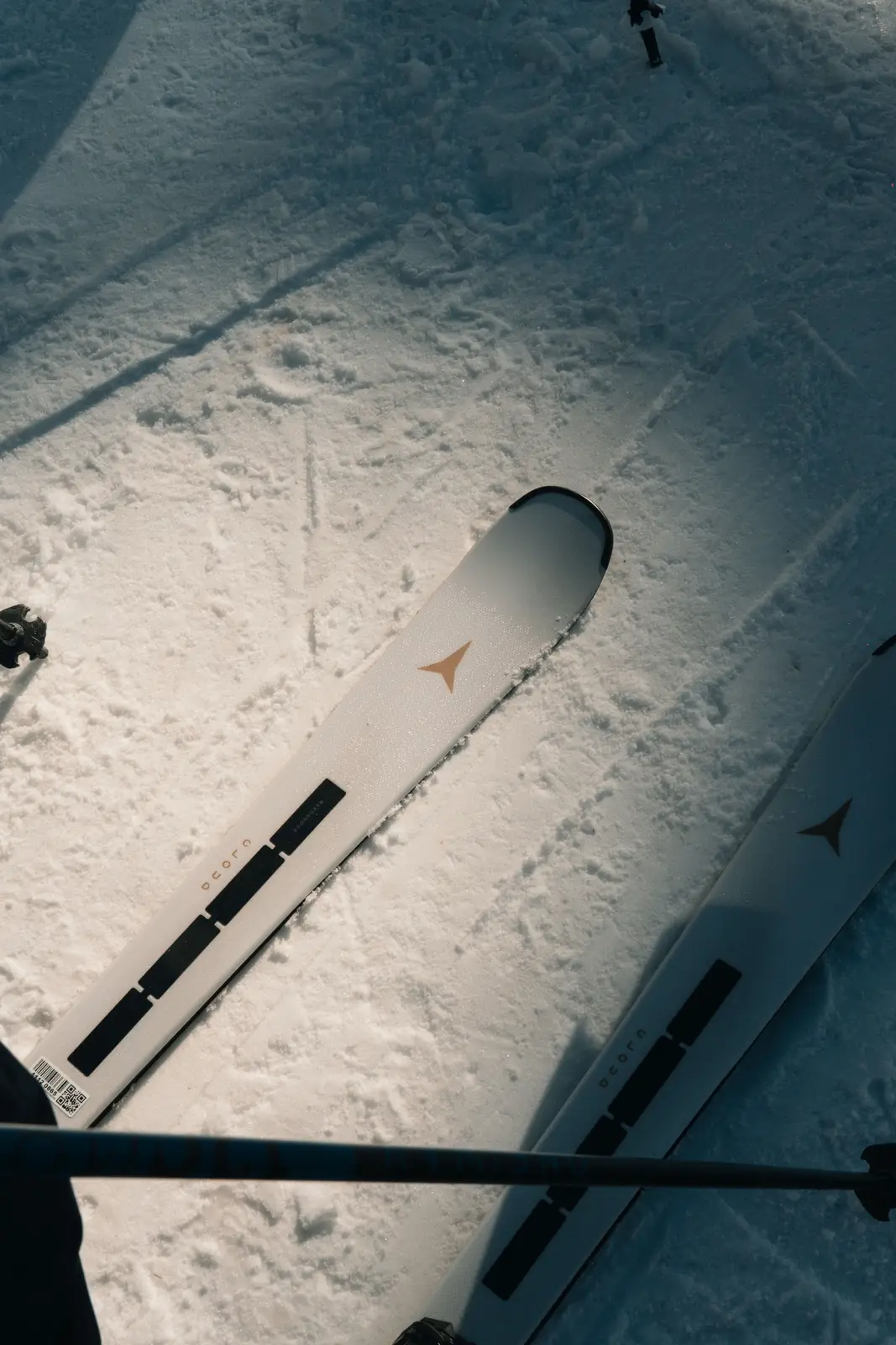 Vue aérienne de skis blancs avec un logo triangulaire doré posés sur la neige, avec un bâton de ski traversant l'image horizontalement.