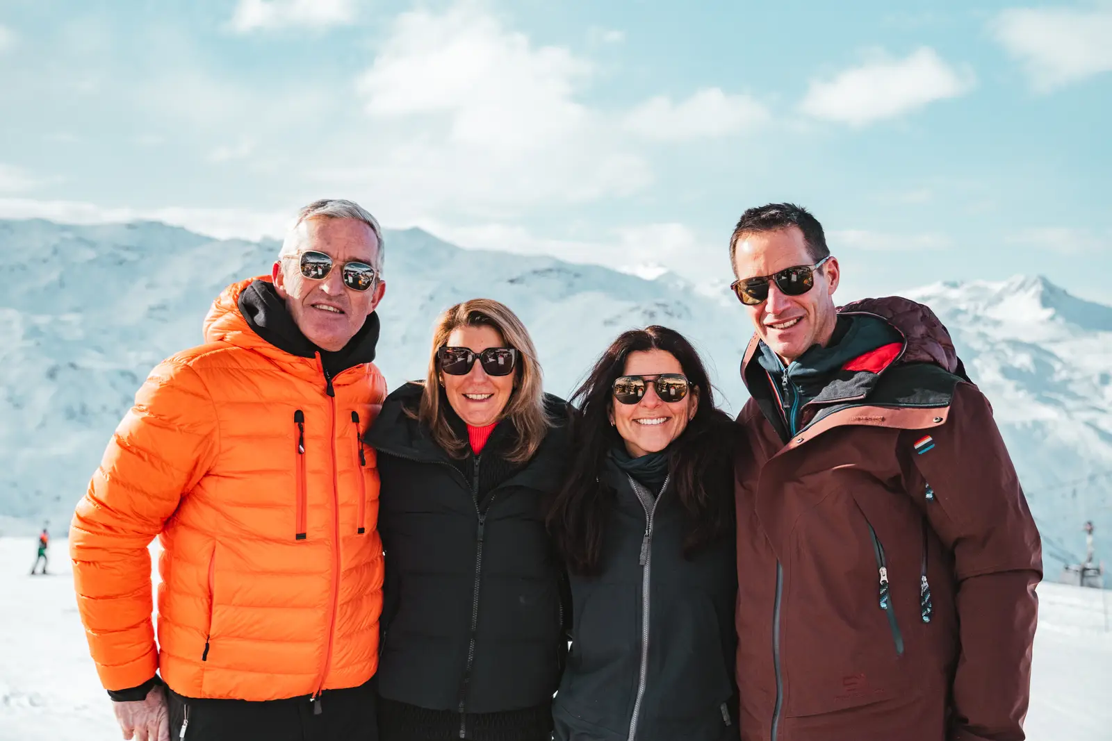 Quatre adultes portant des vestes d'hiver et des lunettes de soleil souriants posent ensemble dans un paysage enneigé de montagnes sous un ciel bleu partiellement nuageux.