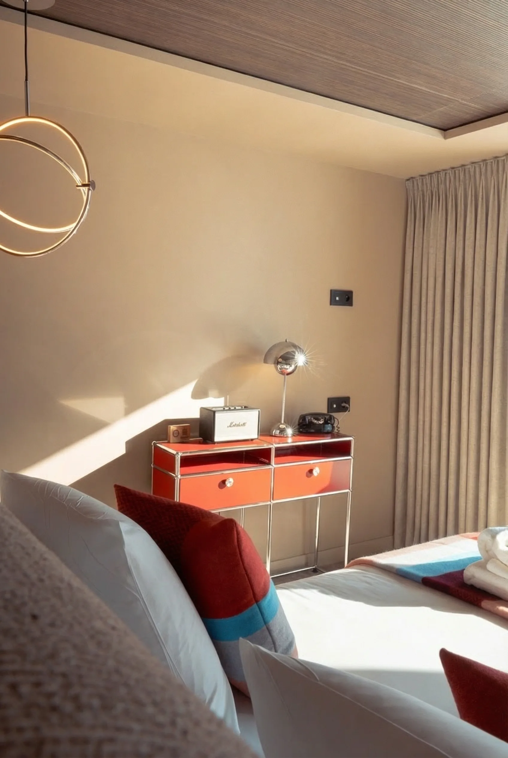 Coin chambre avec lit partiel sur lequel repose un oreiller rouge à rayures bleues, une table de chevet rouge à deux tiroirs avec une lampe chromée, un radio vintage et un téléphone noir, lumière naturelle entrant par la fenêtre à droite avec un rideau beige.