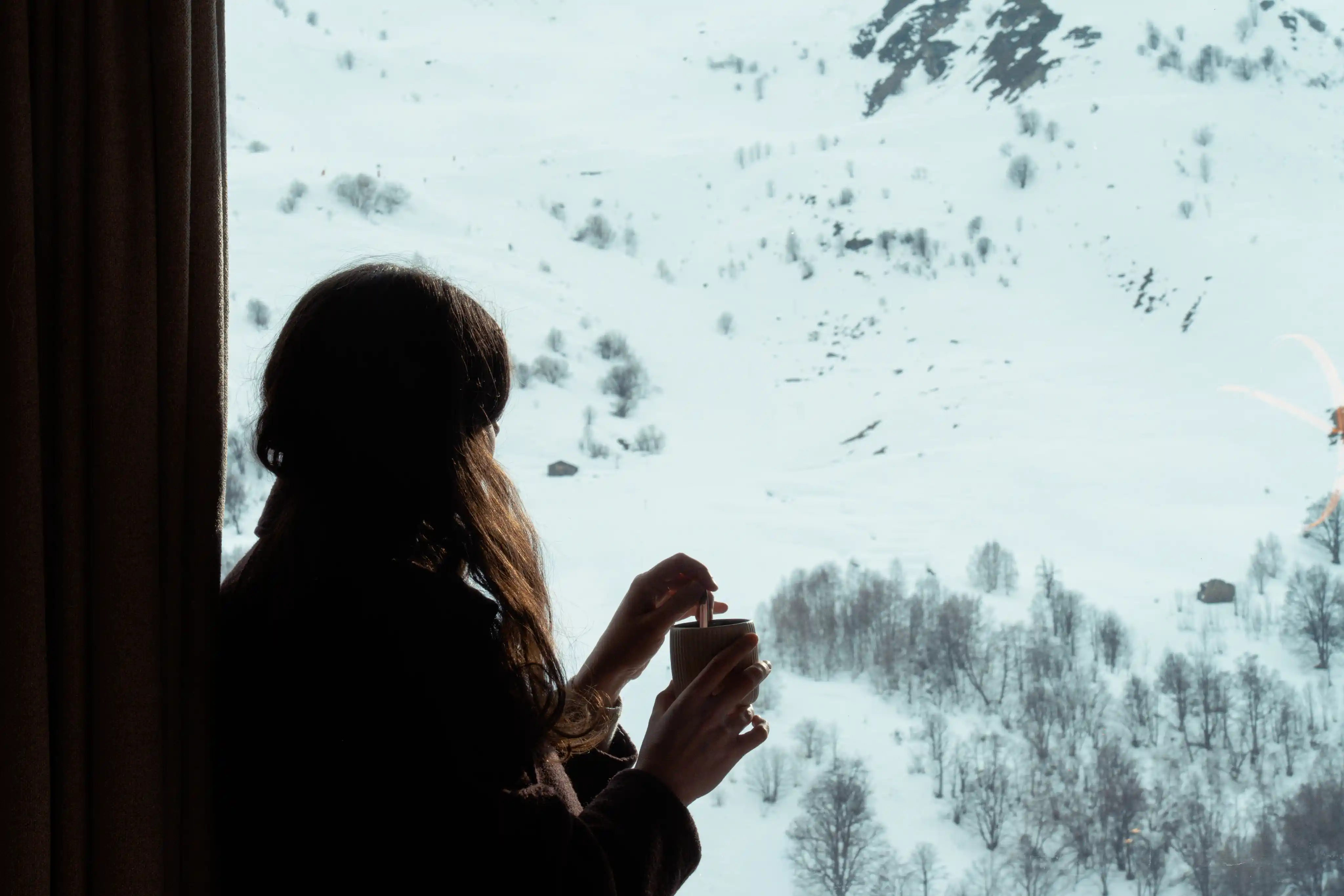 Une femme aux cheveux longs vue de dos, tient une tasse près d'une fenêtre avec vue sur un paysage enneigé de montagne et d'arbres.