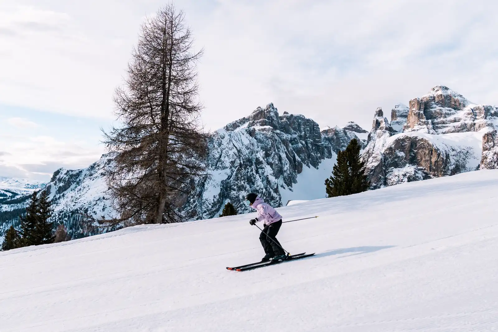 Une personne en tenue de ski violette descend une pente enneigée avec des montagnes rocheuses enneigées en arrière-plan et des arbres dispersés.