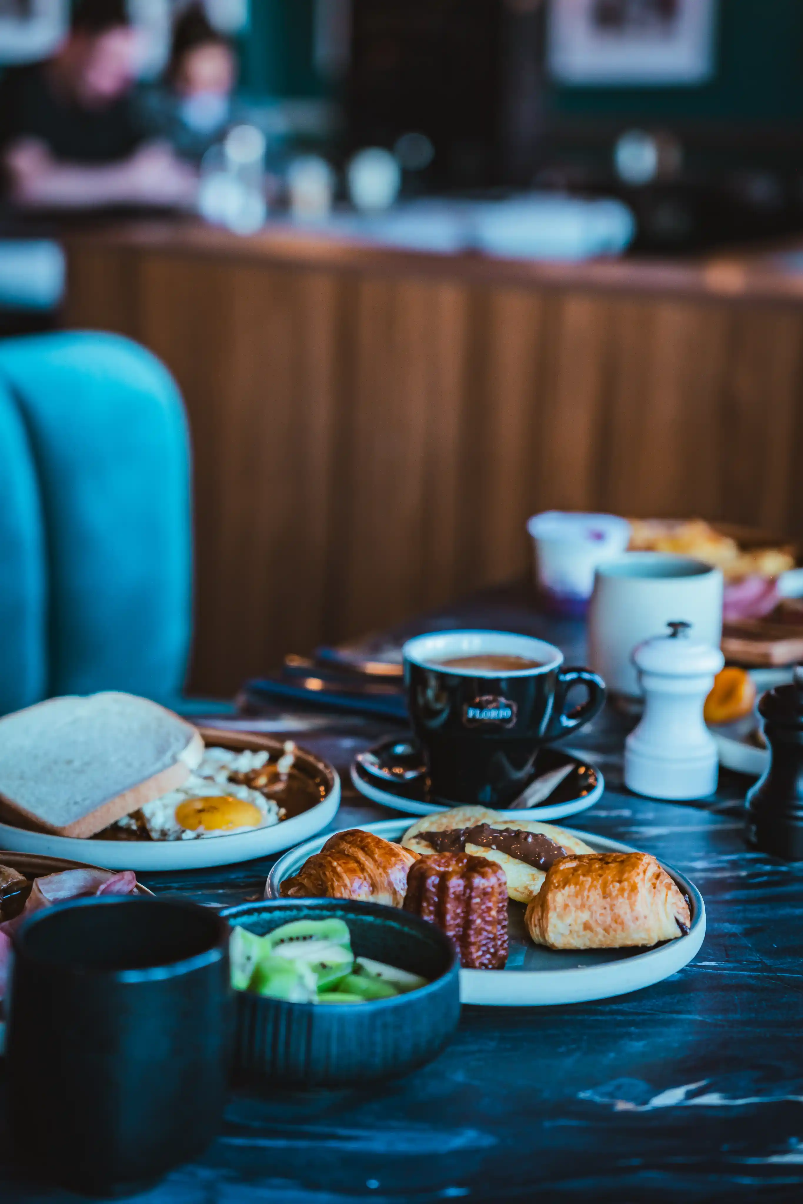 Petit déjeuner sur une table avec café dans une tasse noire, un œuf au plat avec deux tranches de pain, assortiments de viennoiseries dont croissants et cannelé, et un bol contenant des tranches de kiwi.